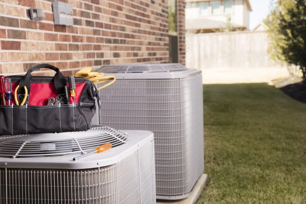 Outdoor view of HVAC units next to a brick house, with a repair tool bag on top of one unit