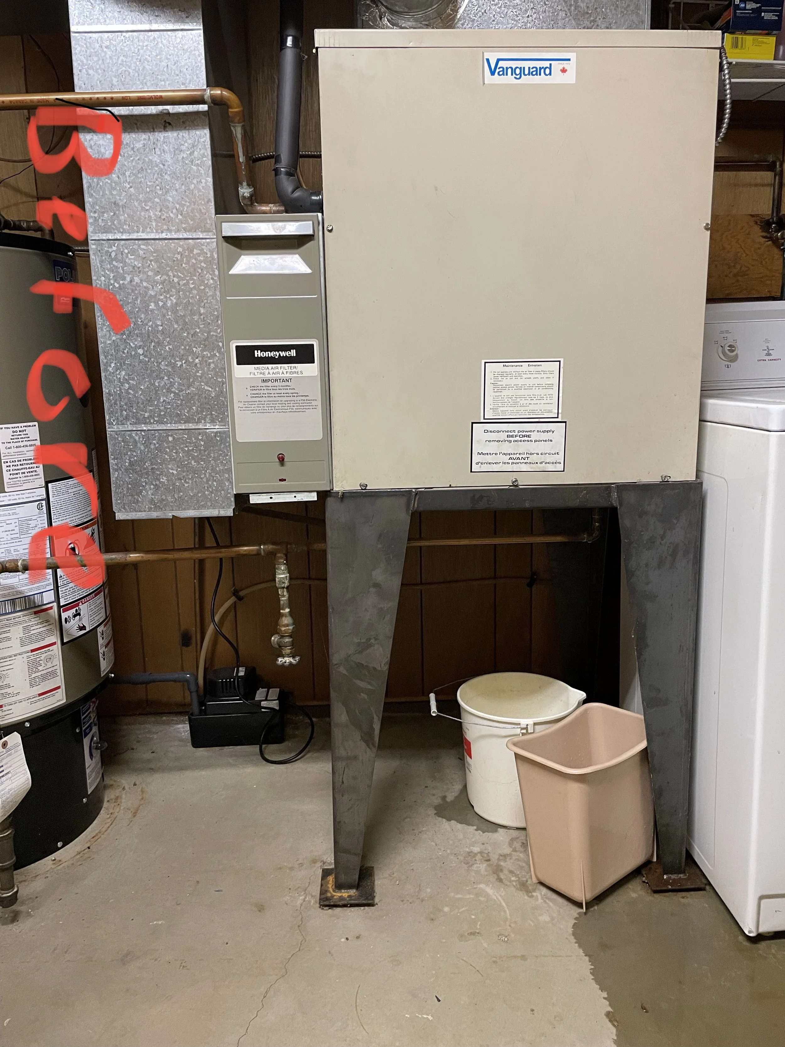 An HVAC unit labeled Vanguard in a laundry room, with a white bucket and a tan trash bin underneath, and a washing machine on the right.