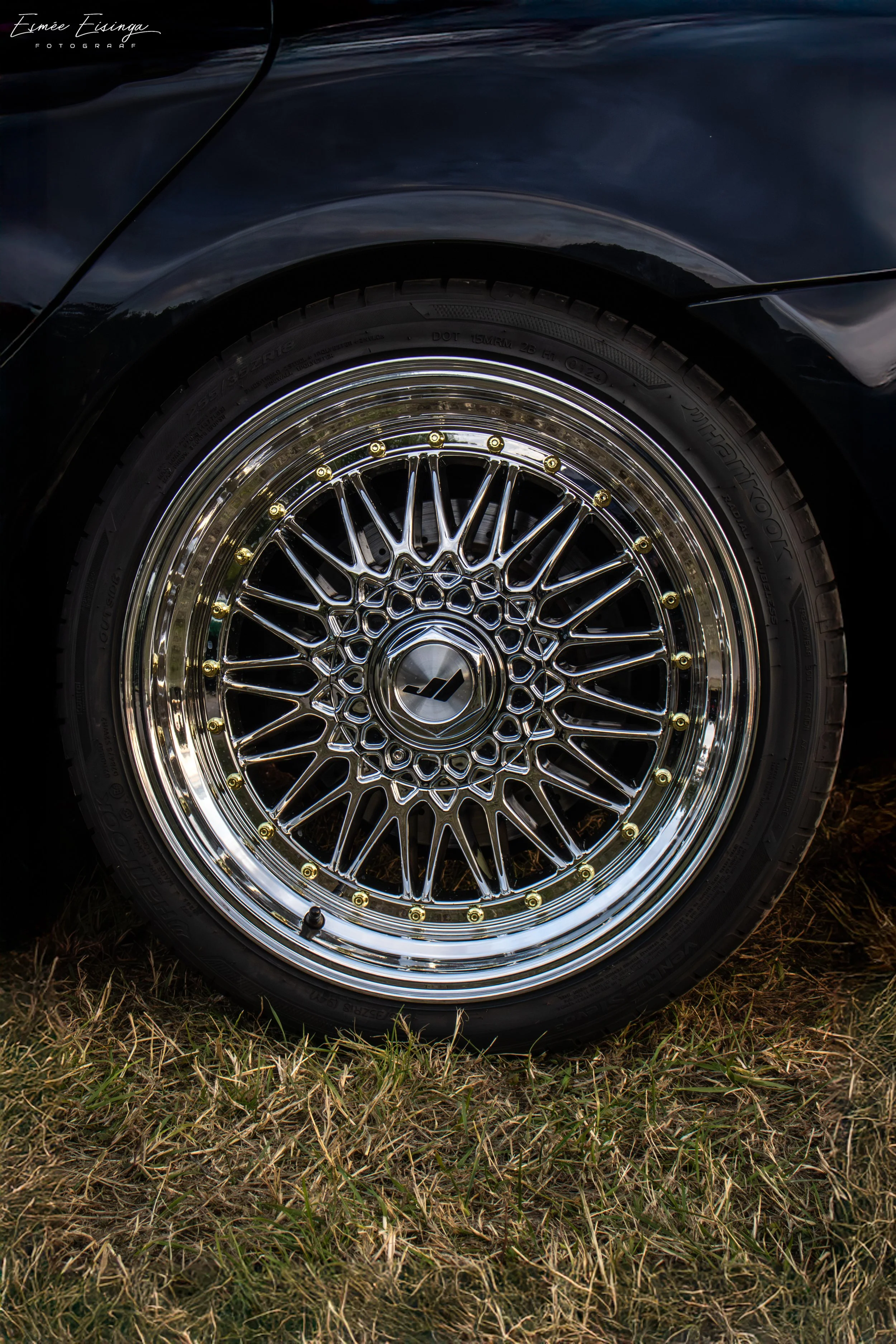Close-up van een zwarte auto met een glanzende gedeukde velg en een profielband, geparkeerd op gras.