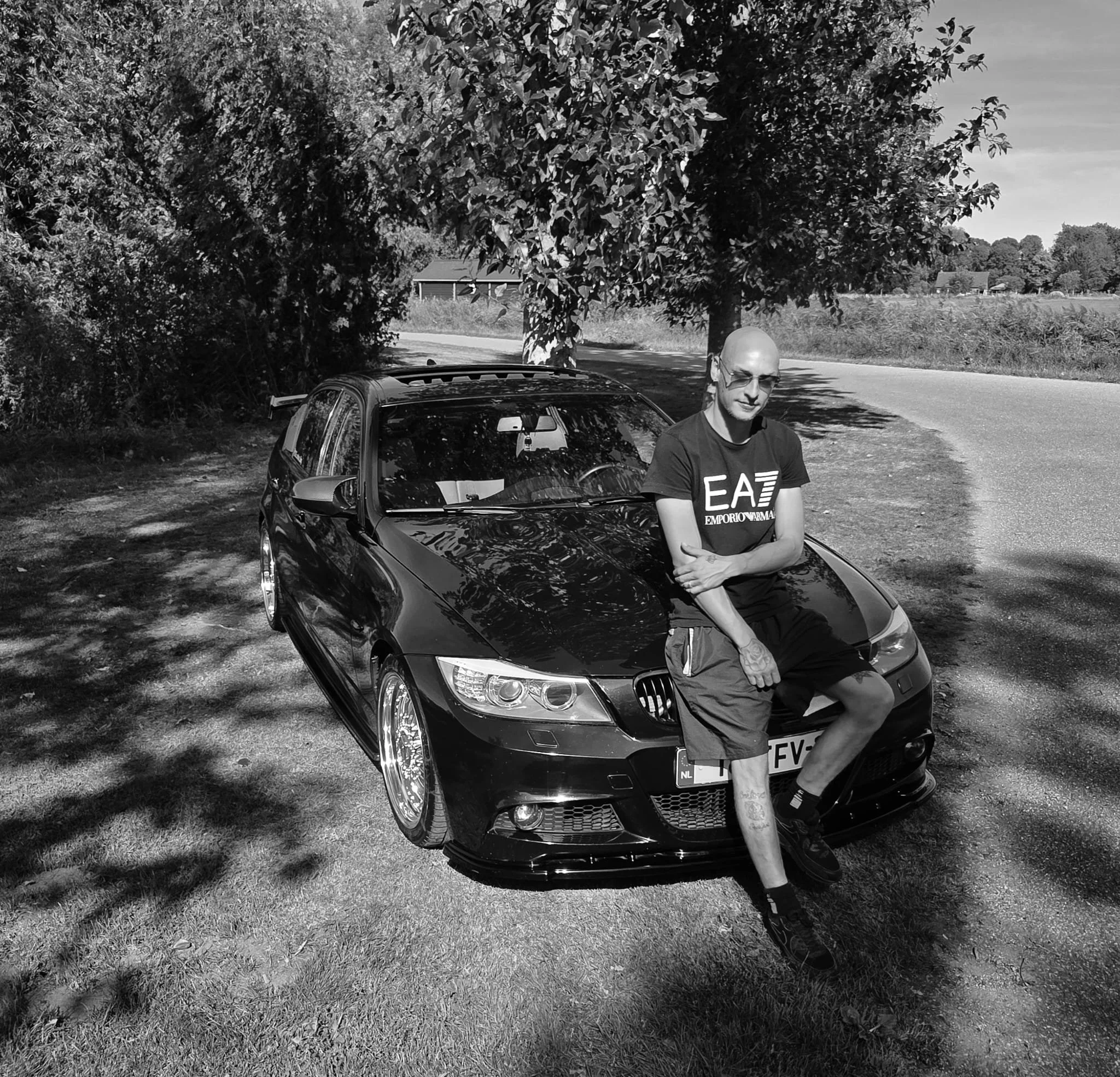 Man met een zonnebril zittend op de motorkap van een zwarte sportwagen, onder een paar bomen langs de weg, in een landelijke omgeving.