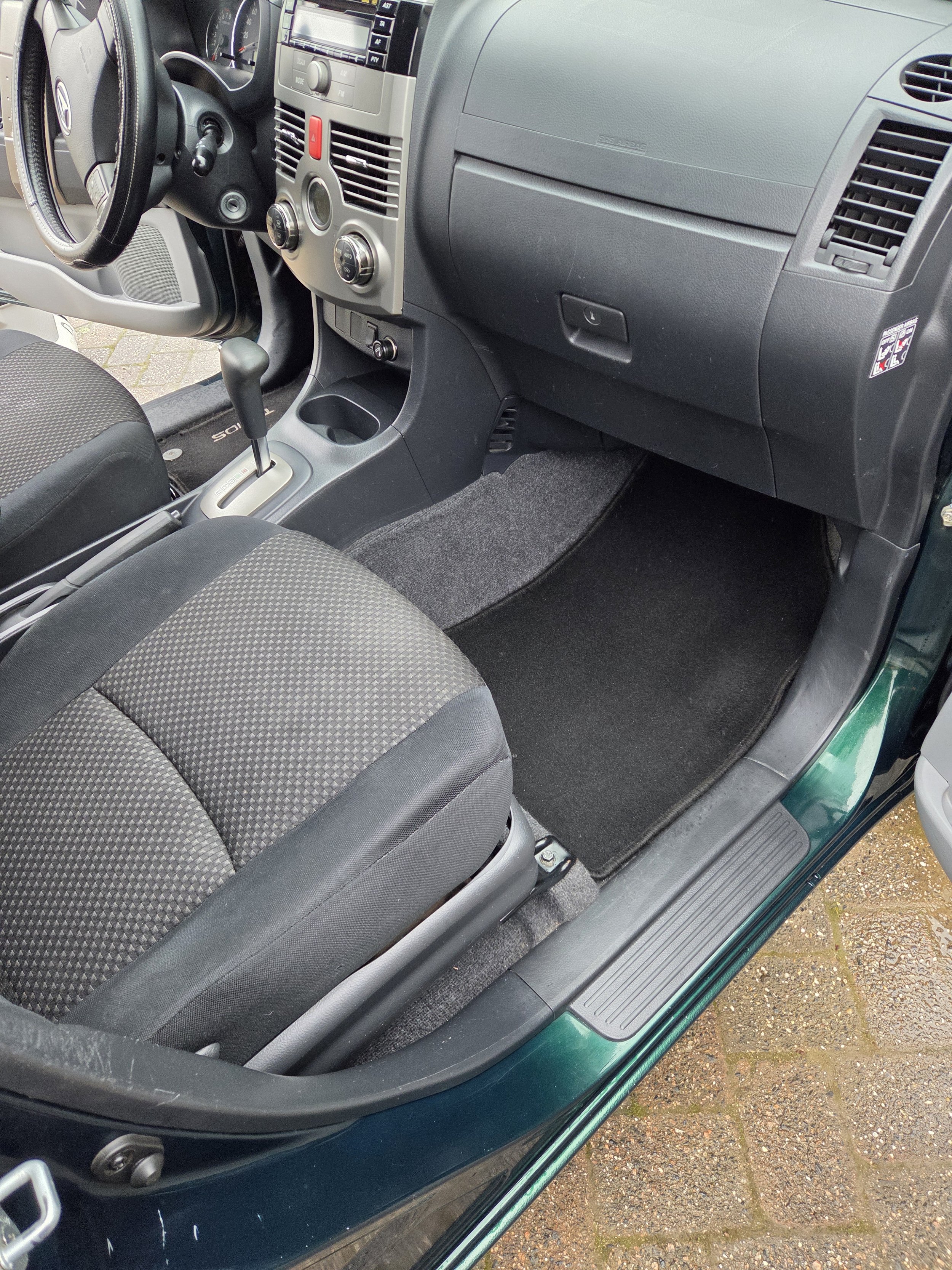 Interieur van een auto, inclusief het dashboard, stuur, versnellingspook en voorstoel. De vloerbedekking en deurrand zijn duidelijk zichtbaar.