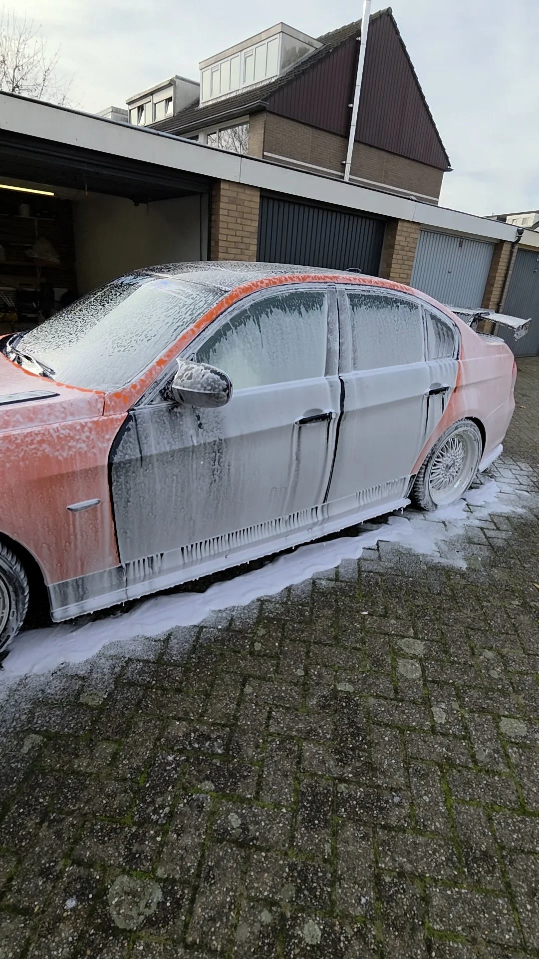 Een auto wordt gewassen en schuimend met zeep bedekt, geparkeerd op een oprit met een garage en huizen op de achtergrond.