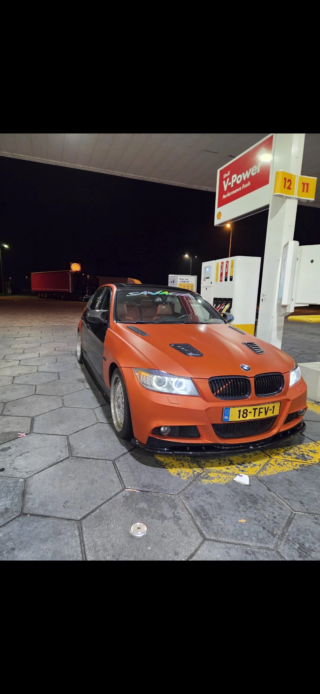Een oranje en zwart gemodificeerde BMW sportauto bij een tankstation 'Shell V-Power' 'Performance Fuels' in Nederland, nacht.