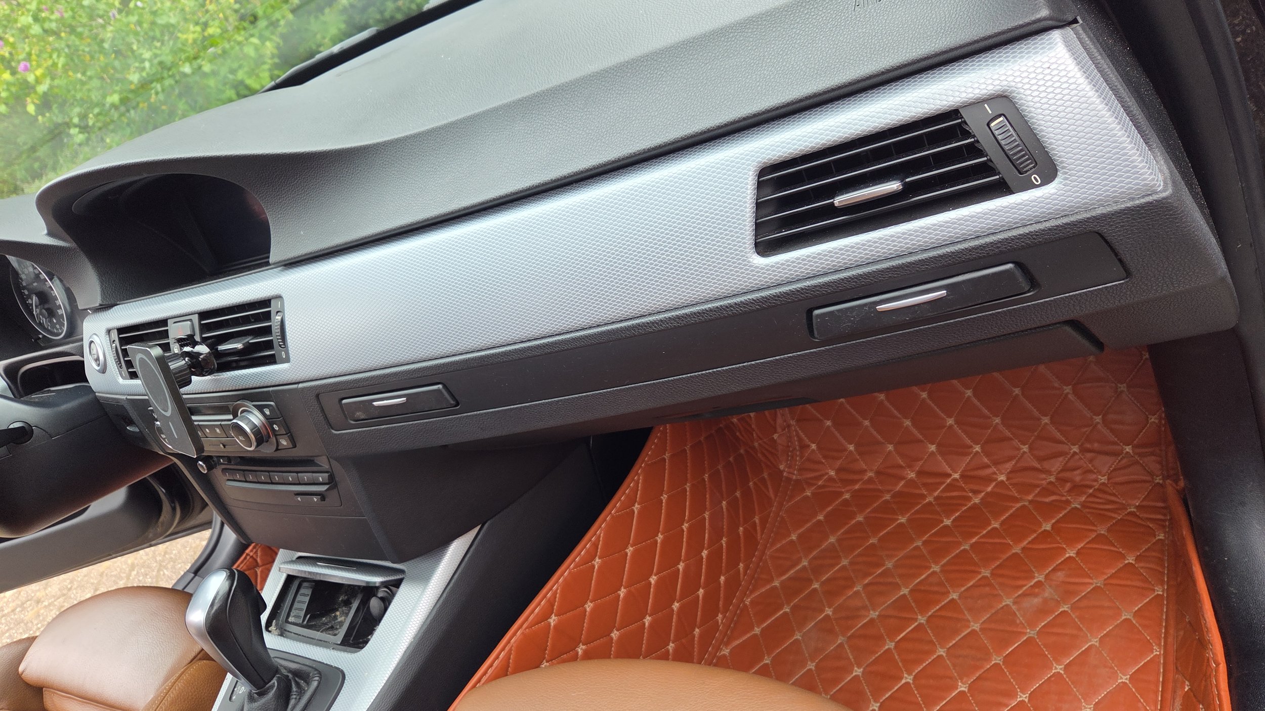 Interieur van een auto met een dashboard, zijpaneel en een lichtbruine stoel, met een quilted orange mat op de vloer.