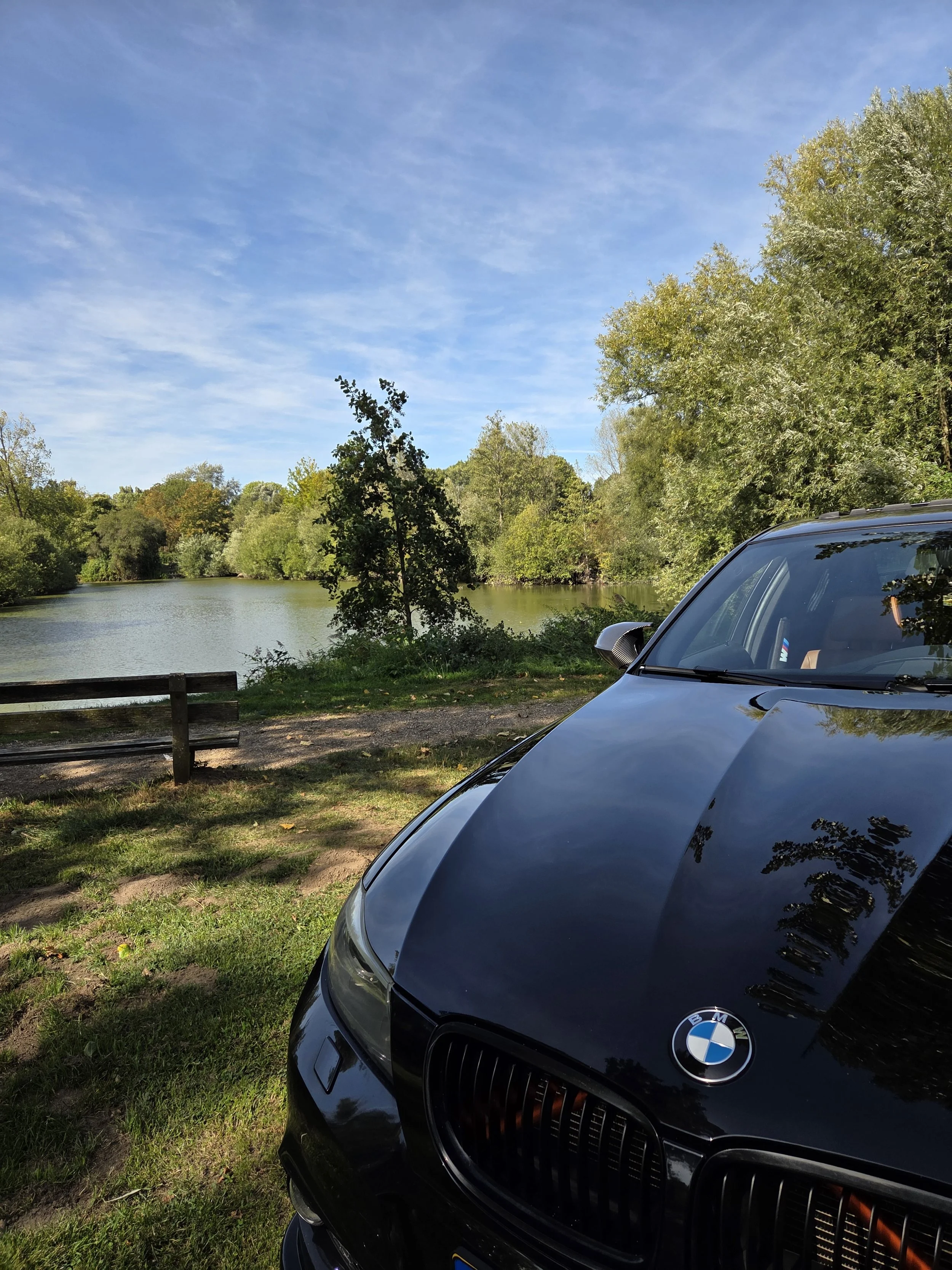 Een zwart BMW auto geparkeerd naast een bosrijk meer. De lucht is blauw met wat lichte wolken.