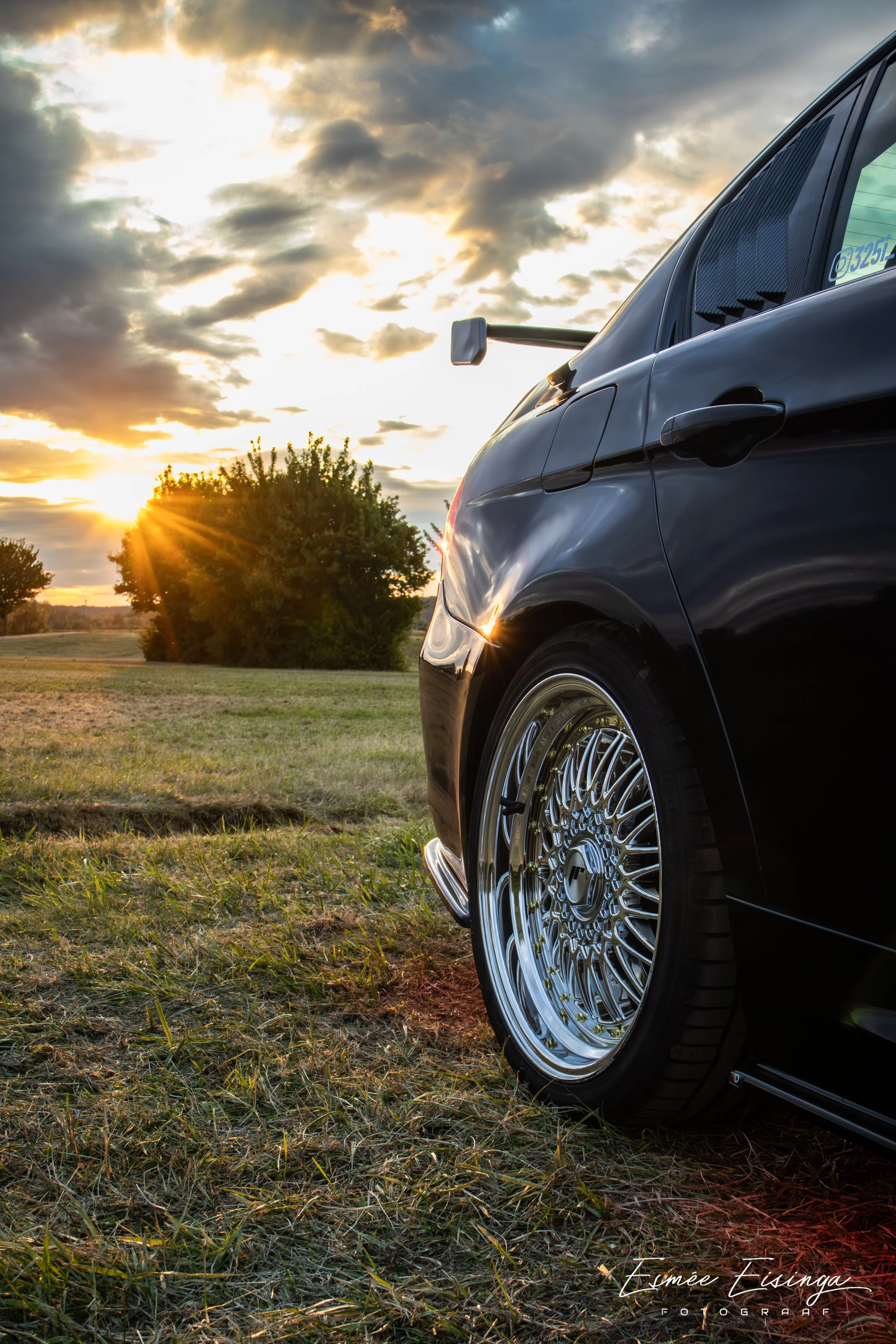 Zwarte sportauto met glanzende velgen in een grasveld bij zonsondergang, met een grote boom en bewolkte hemel op de achtergrond.