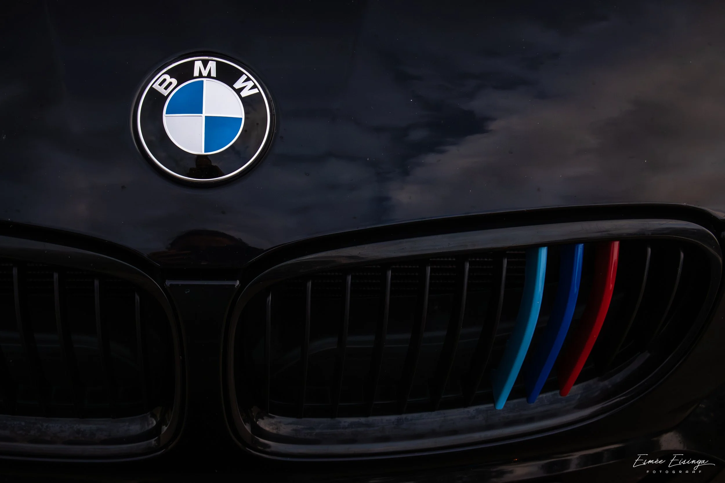 Close-up van de voorkant van een zwart BMW auto, met de BMW-logo en een grille met drie gekleurde accenten in blauw, donkerblauw en rood.