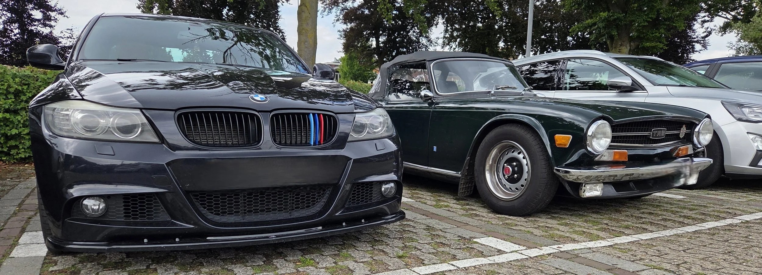 Vijf auto's geparkeerd naast een boom, waaronder een moderne zwarte BMW en een klassieke groene auto.