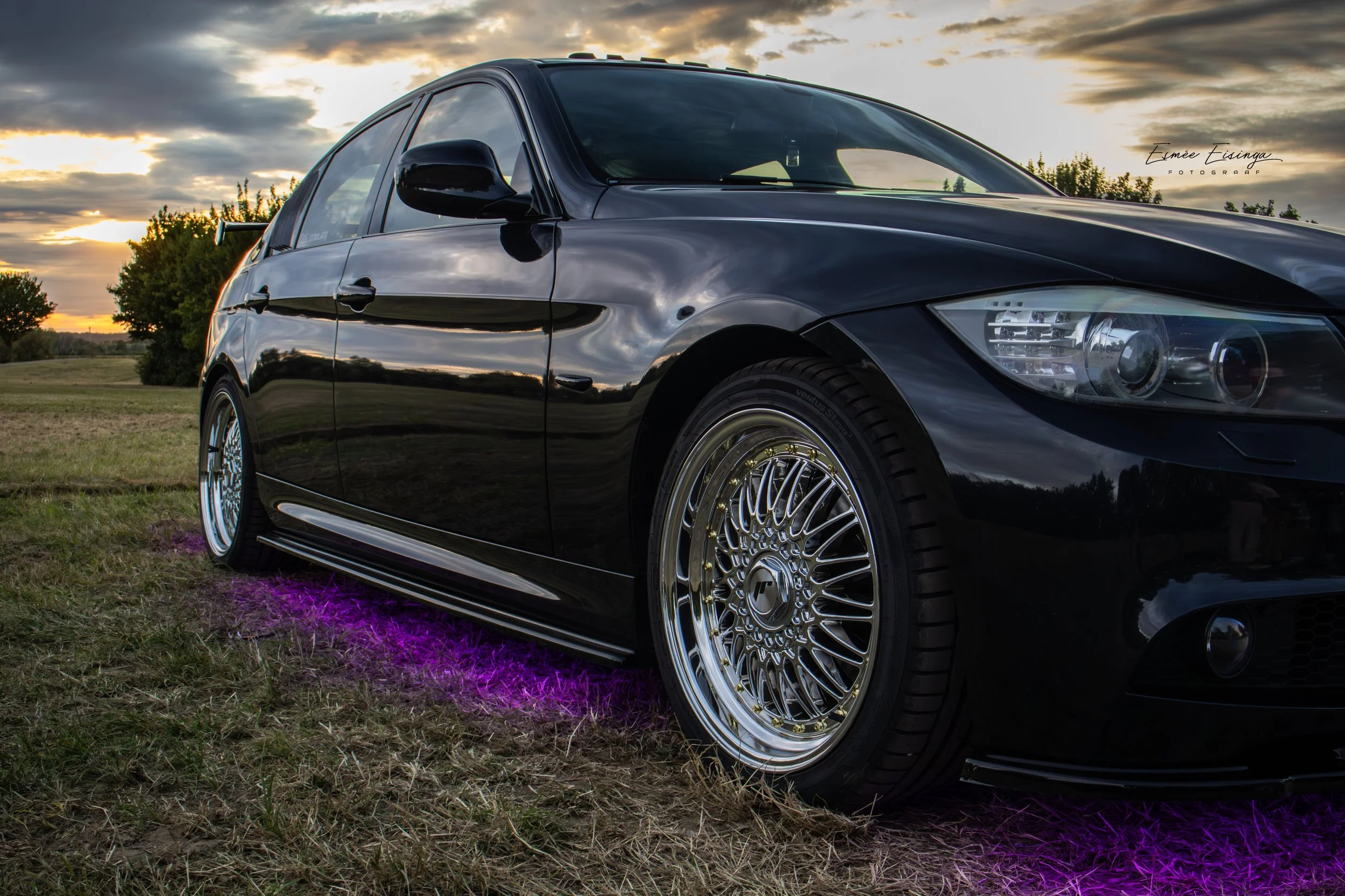 Zwarte sportwagen met gedraaide velgen en onderlicht dat paars licht uitstraalt, geparkeerd op grasvelden tijdens zonsondergang.