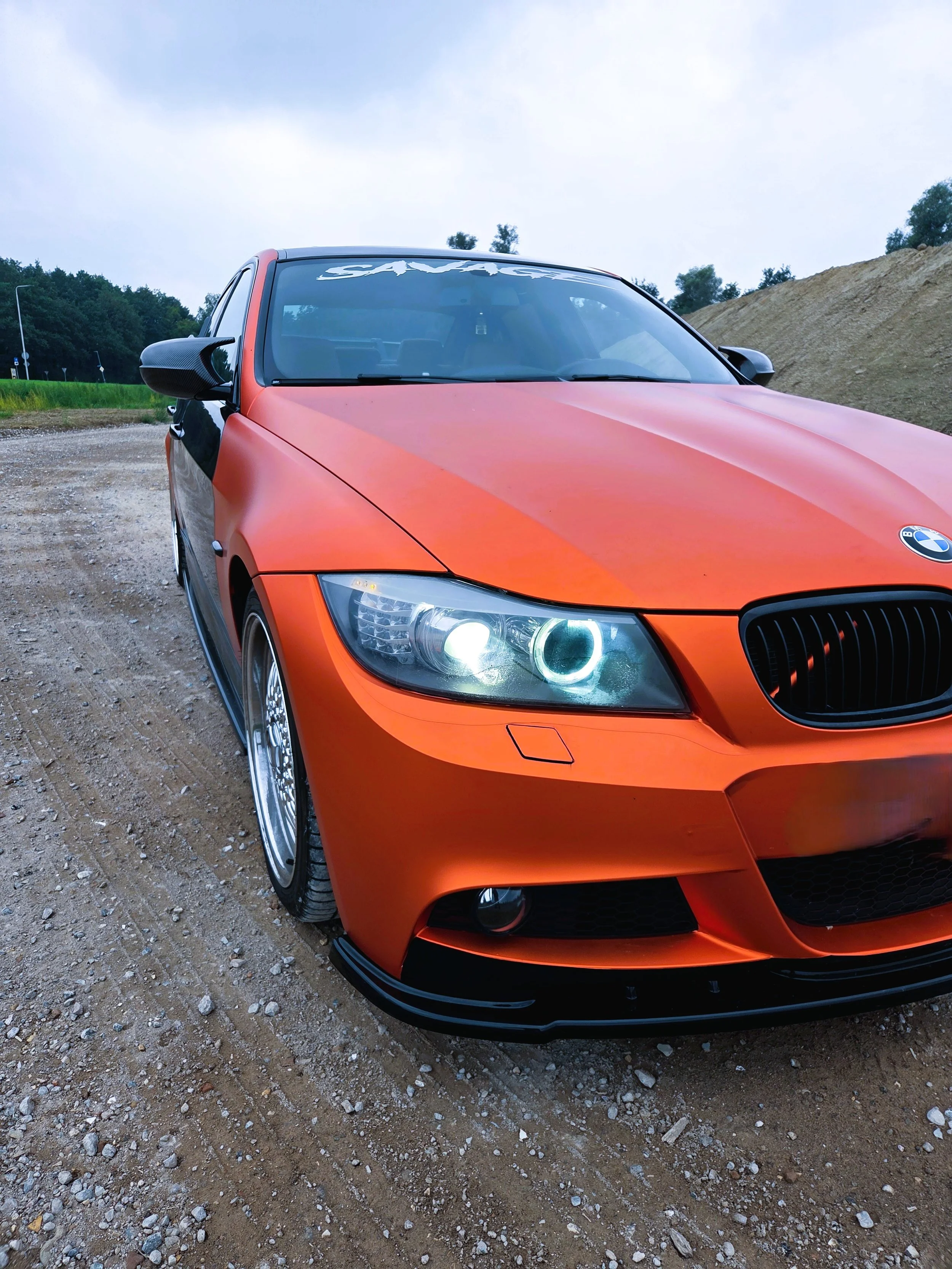 Een sportieve BMW met oranje en zwarte kleur op een gravelweg, met een bos en een hoop aarde op de achtergrond.