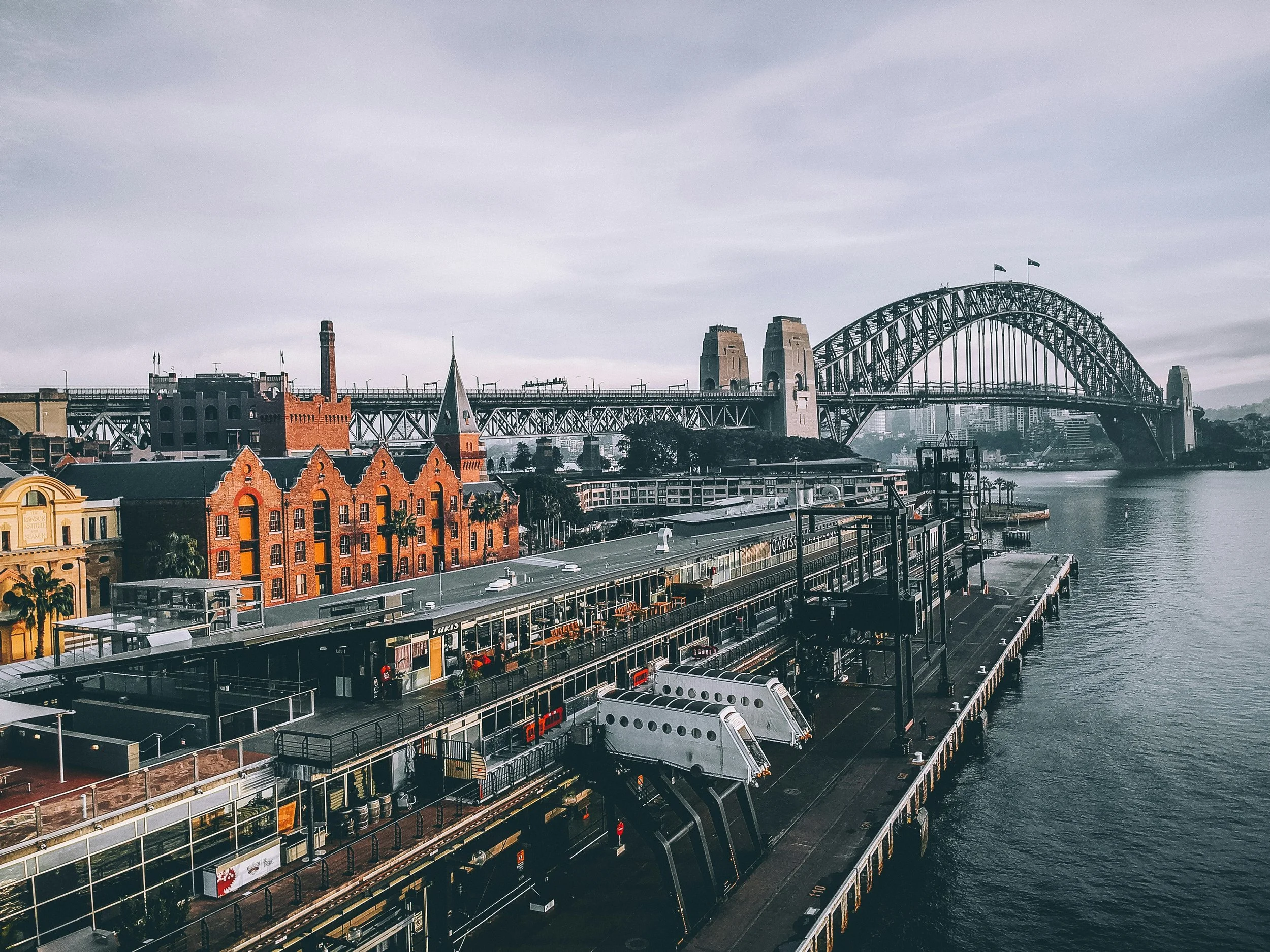View of Sydney Harbour with the Harbour Bridge in the background, a pier and buildings in the foreground, and cloudy skies.