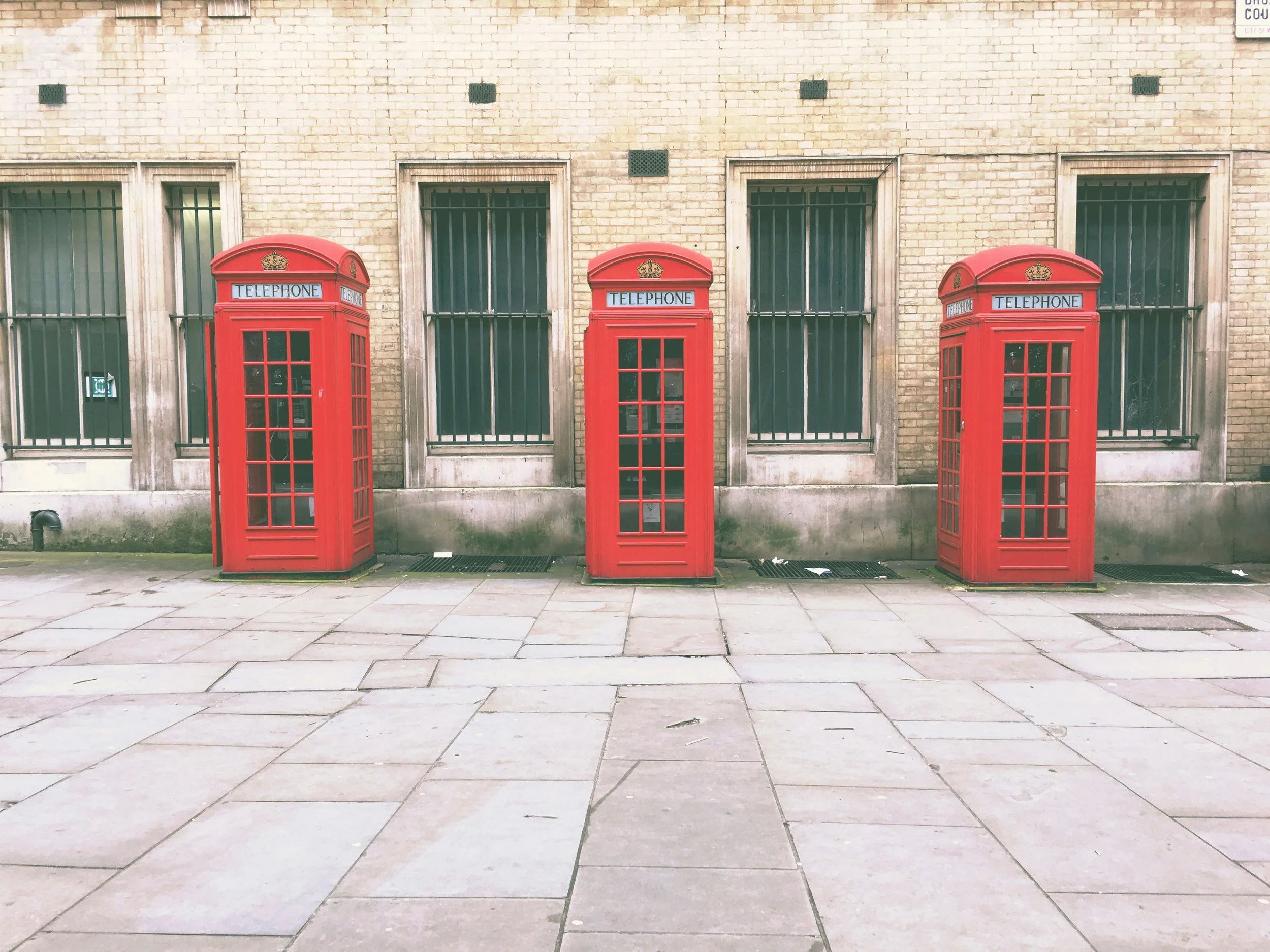 Three red British telephone booths placed on a sidewalk in front of a brick building with barred windows.