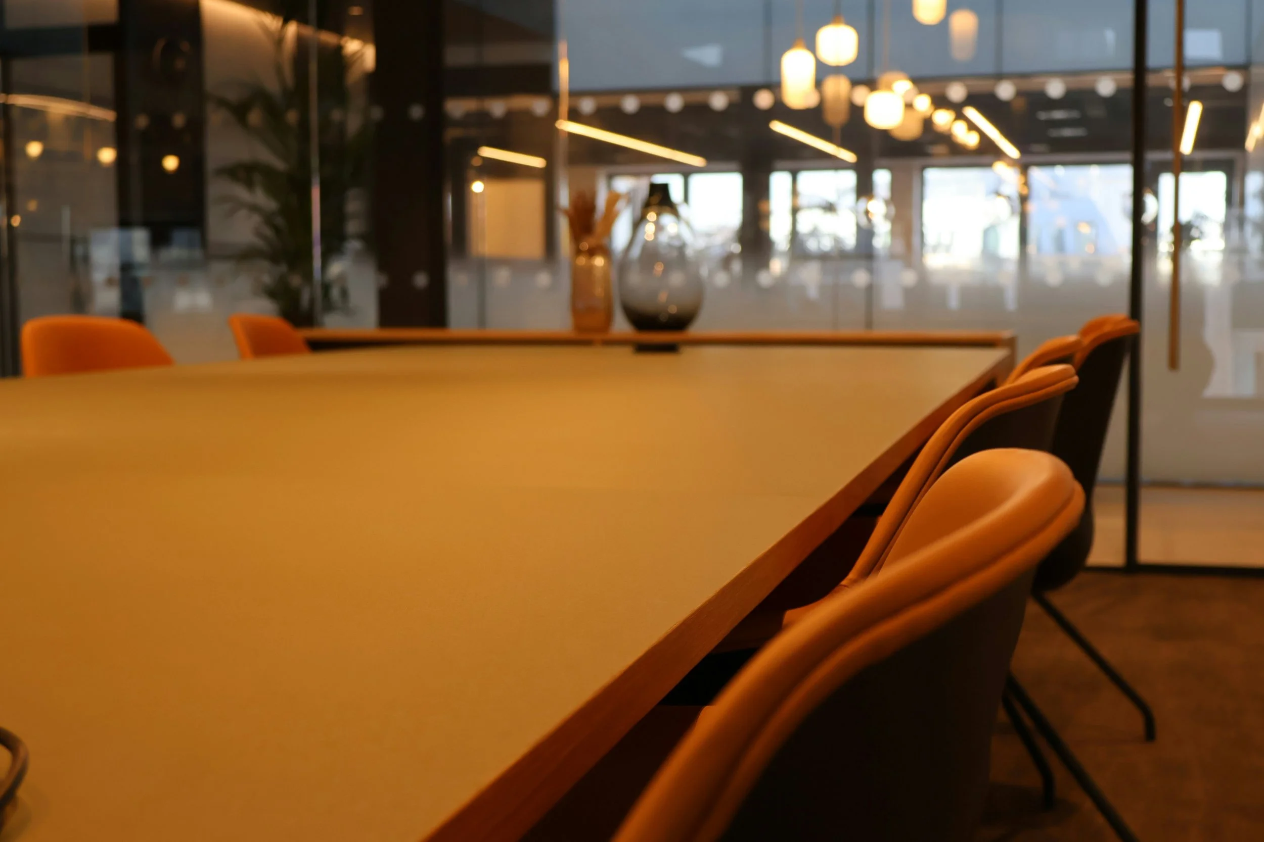 A modern conference room with a long yellow table, orange and black chairs, decorative vases, and contemporary hanging lights. Large glass windows reveal an outdoor view.