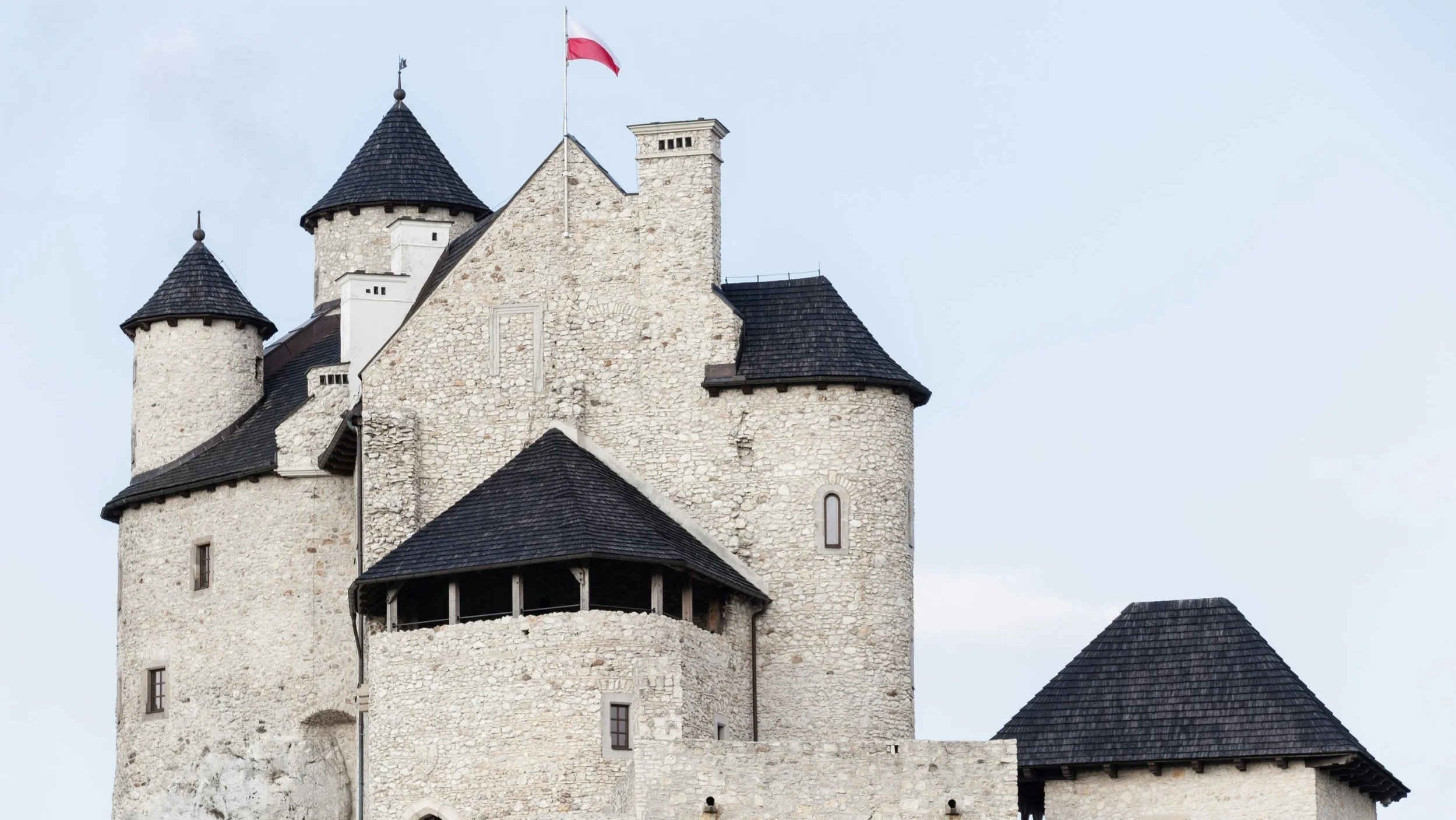 Historic stone castle with multiple towers and dark conical roofs, and a Polish flag on top.