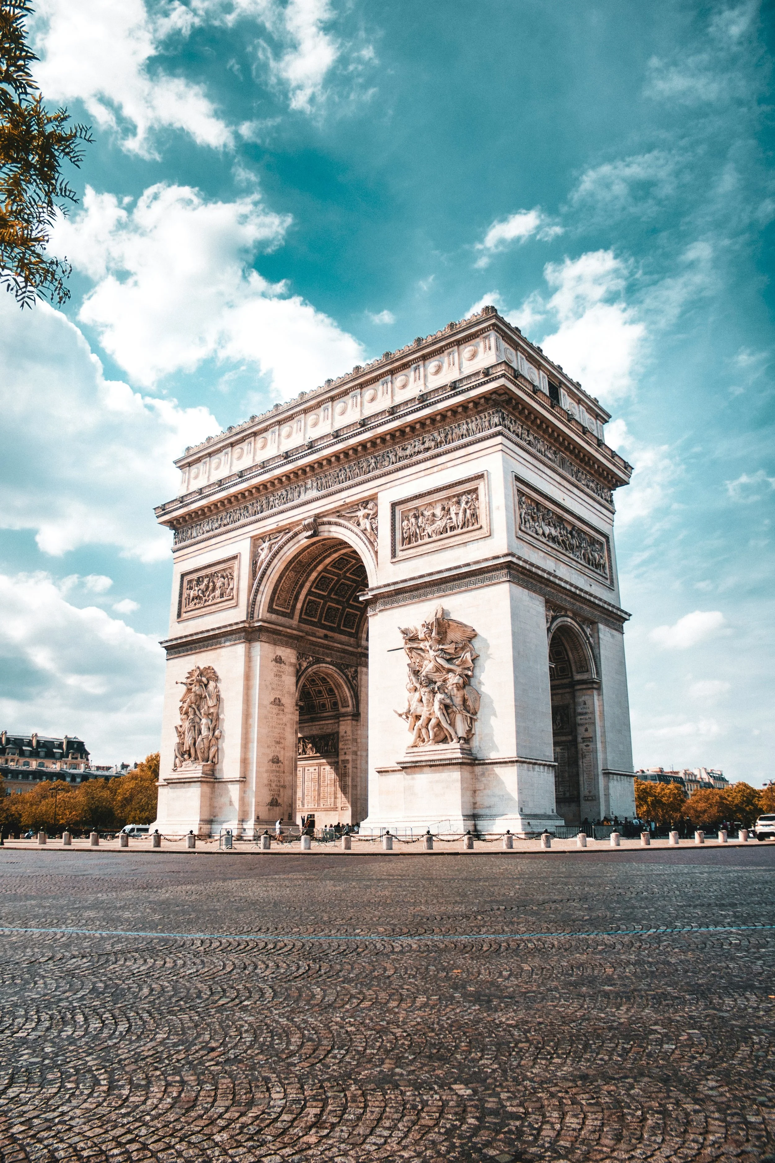 The Arc de Triomphe in Paris, France, with detailed sculptures on its pillars and a cobblestone street in the foreground, under a partly cloudy sky.