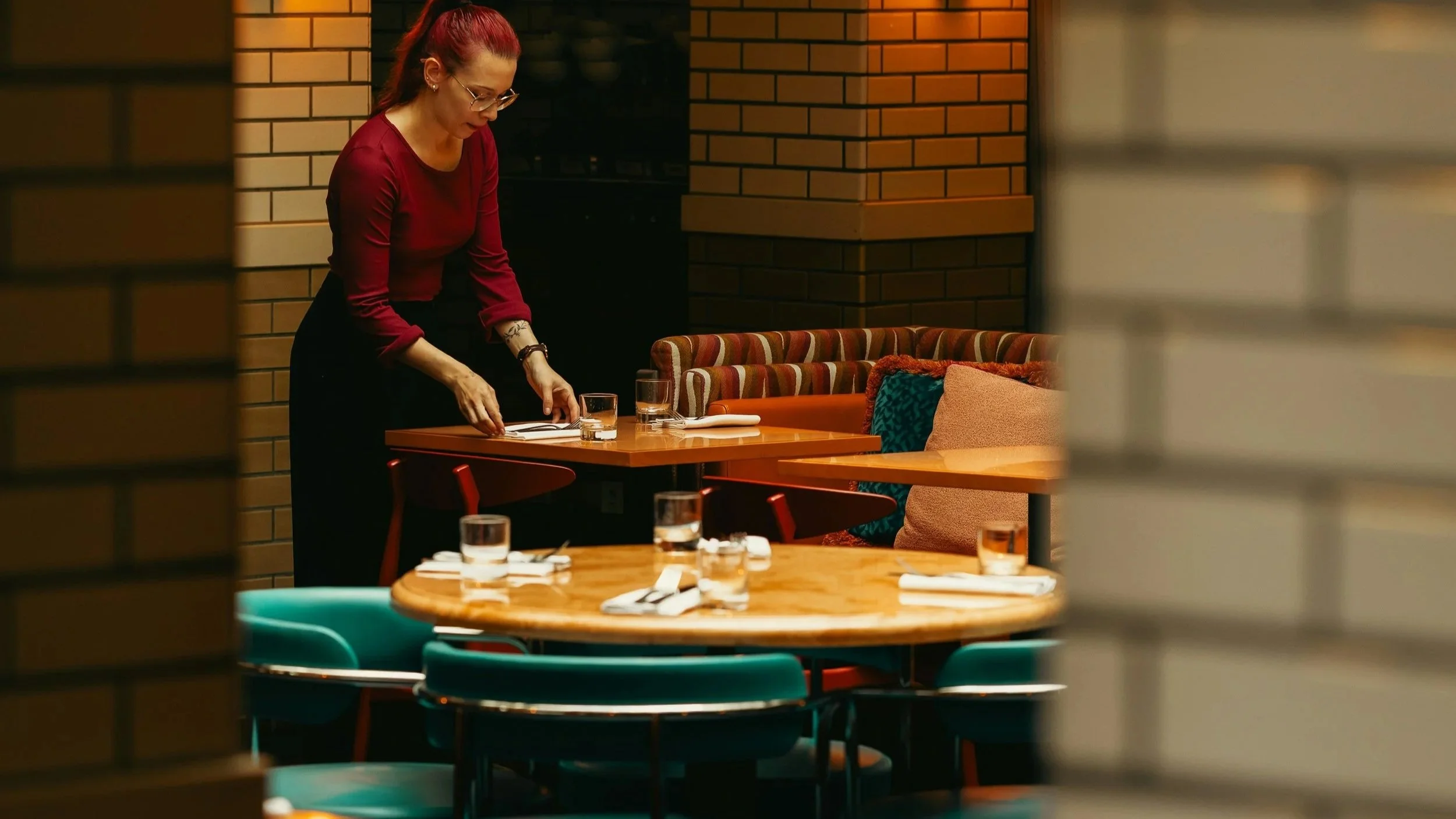 A woman with red hair, glasses, and a red shirt prepares a table in a restaurant, with tables set with glasses and napkins, seen through a partially open window.