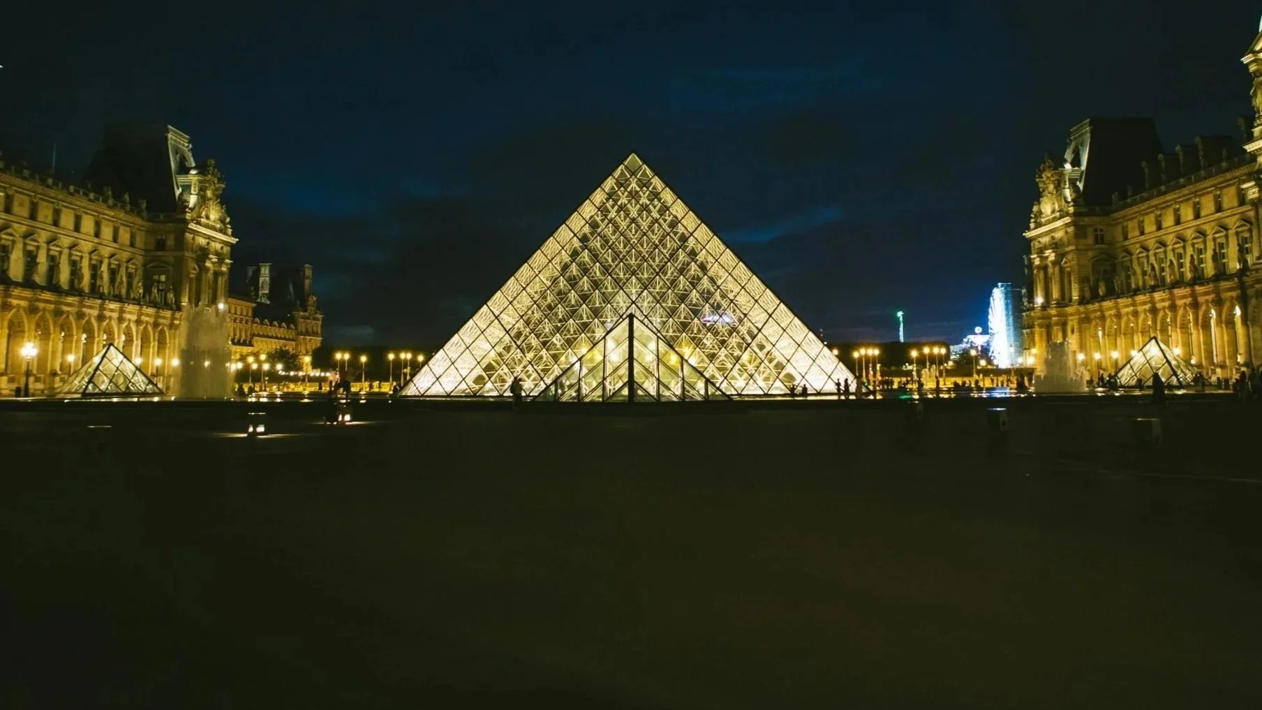 The illuminated glass pyramid of the Louvre Museum at night, surrounded by historic palace buildings with decorative facades and lit windows.