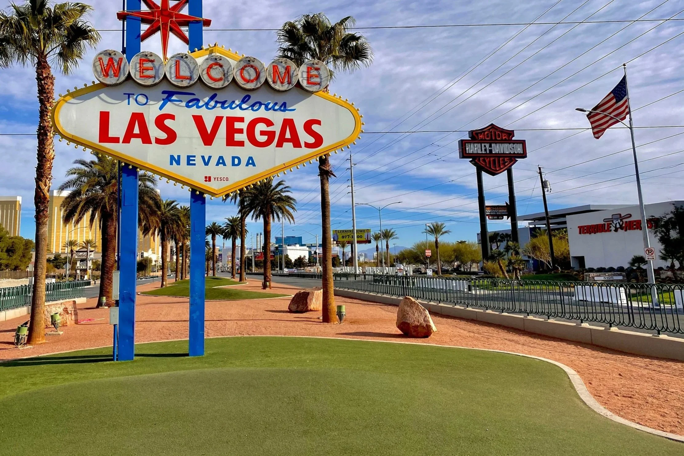 Welcome sign for Las Vegas, Nevada with palm trees in the background, a blue sky with some clouds, and various signs including Harley-Davidson, Terrible Herbst, and an American flag.