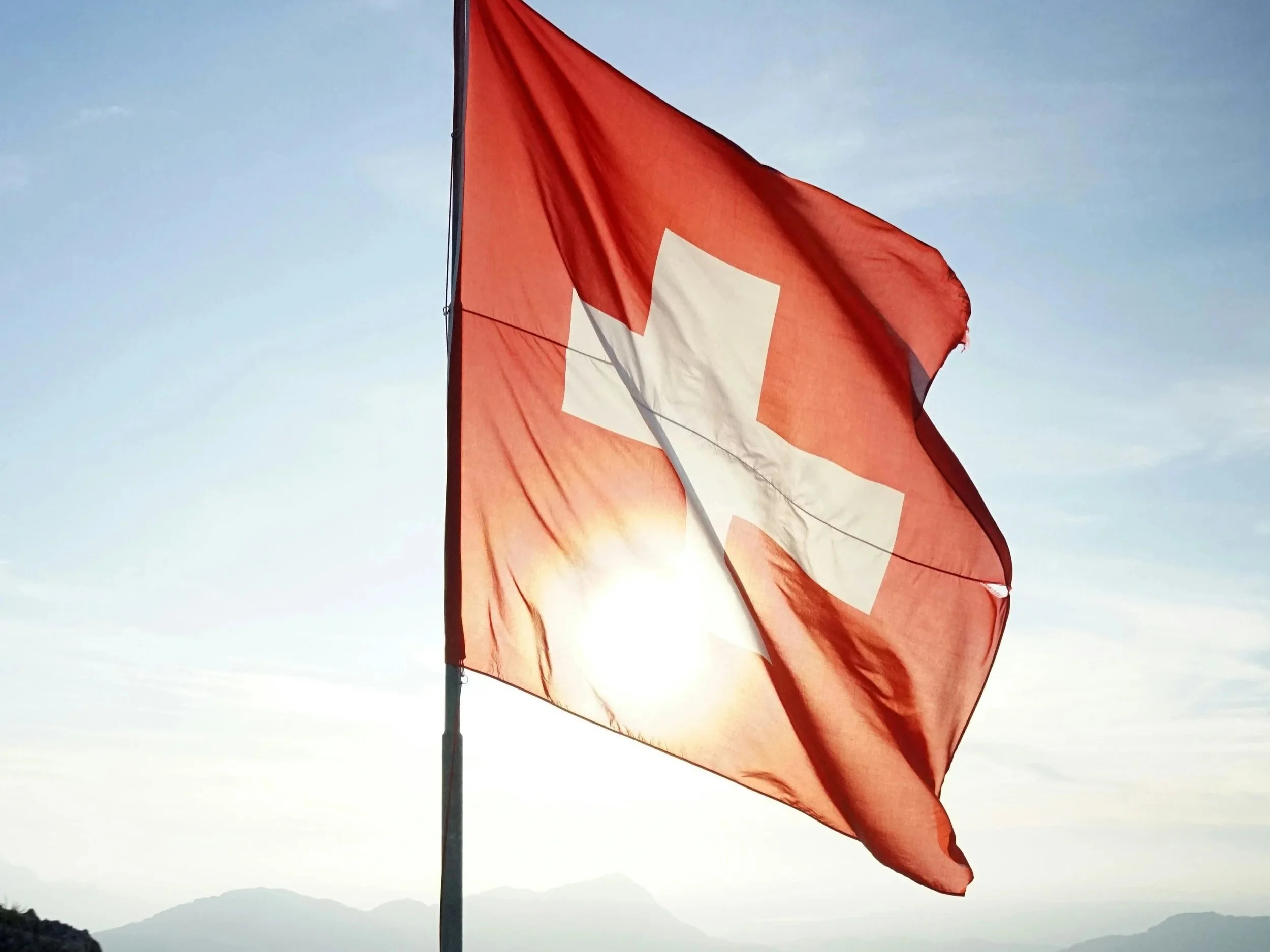 A red flag with a white Swiss cross flutters in the wind during sunset over a mountainous landscape.
