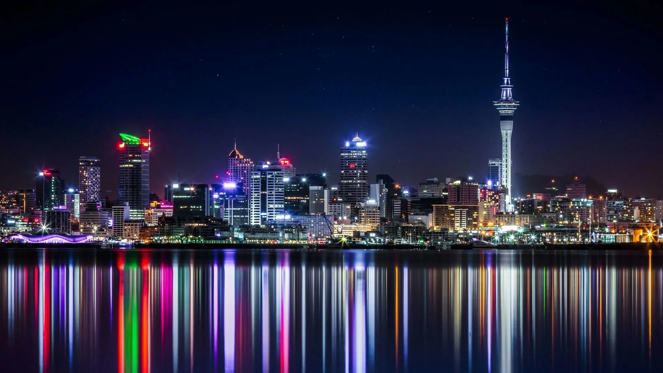 Night view of a city skyline with illuminated buildings, including a tall tower with bright lights, reflected in the water in the foreground.