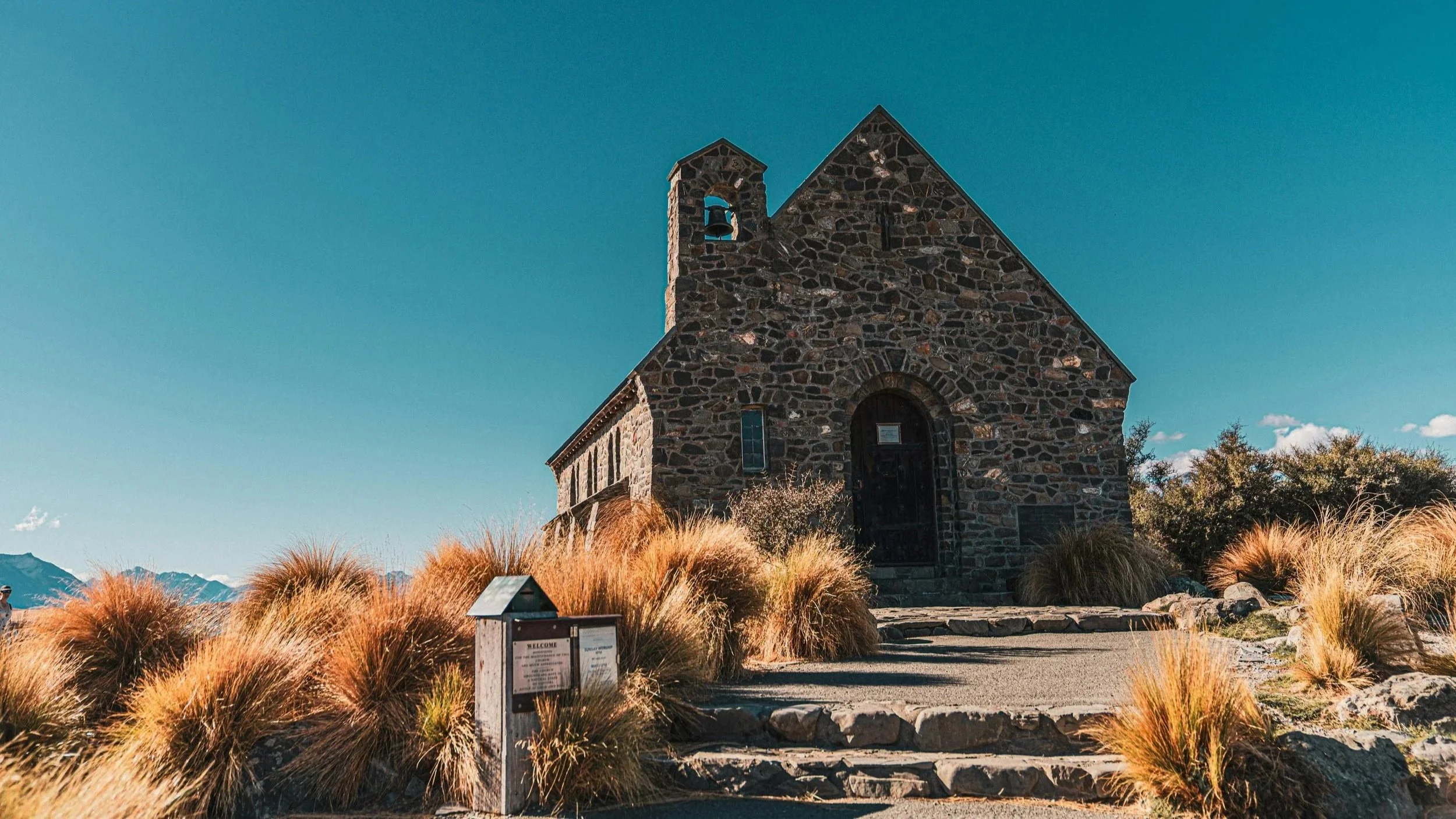 A small stone church with a bell tower, surrounded by dry grass and rocks, under a clear blue sky with mountains in the distance.