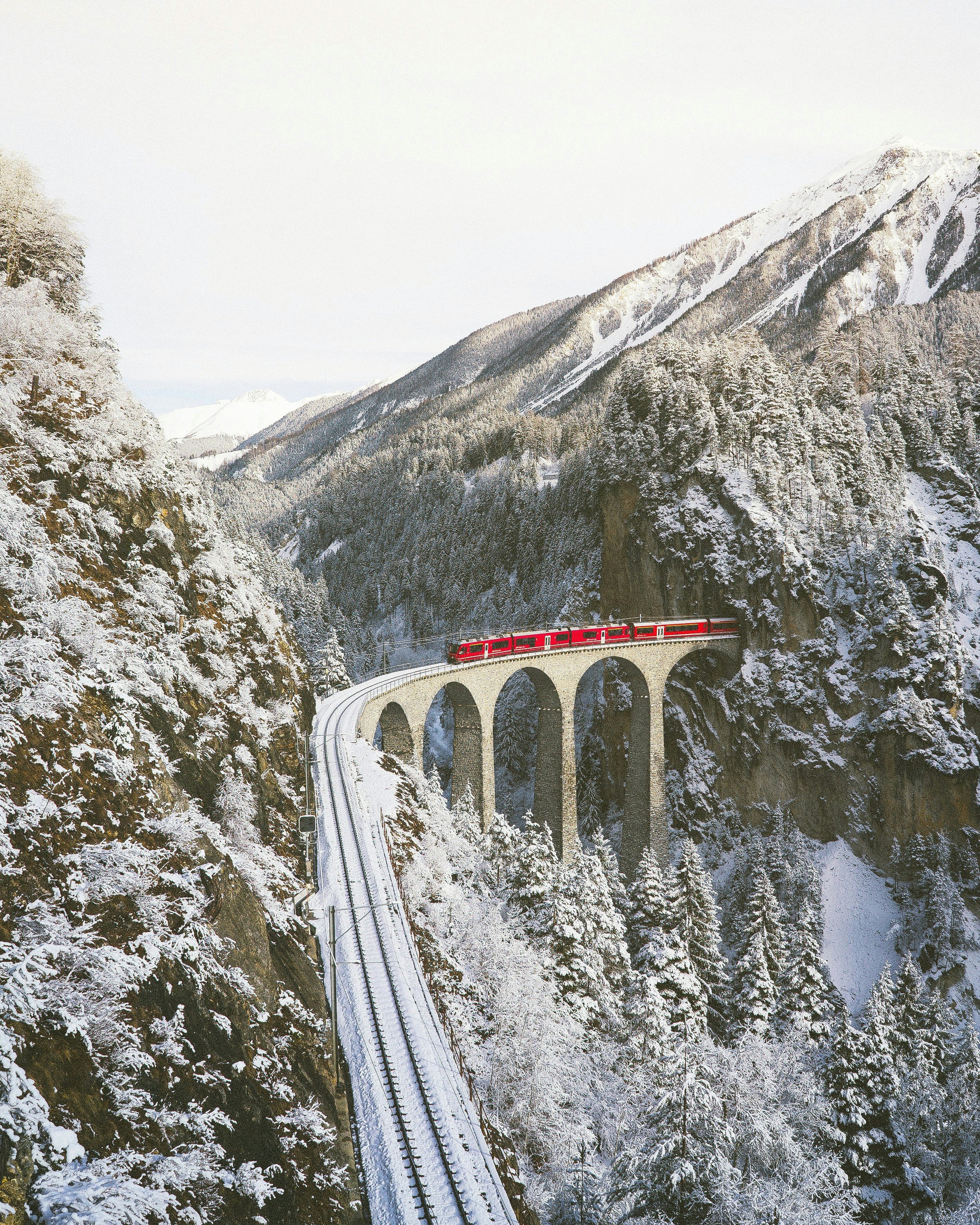 Red train traveling across a snow-covered mountain landscape on a stone arched viaduct.