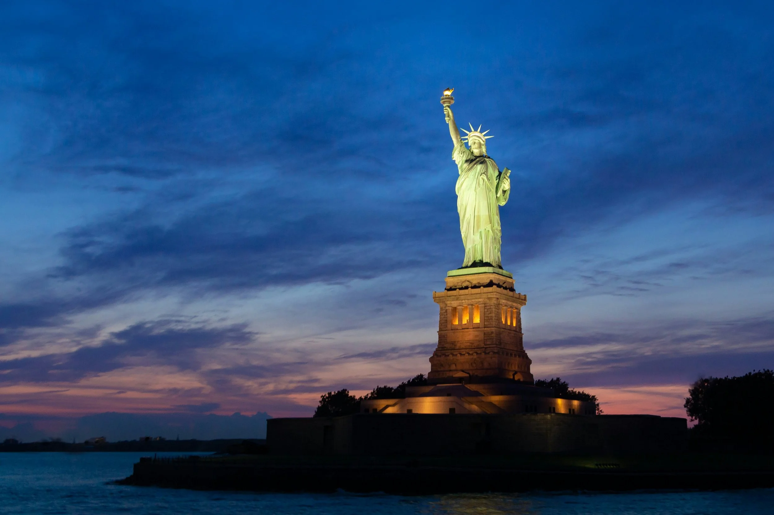 The Statue of Liberty on Liberty Island with an American flag flying in the foreground against a bright blue sky.