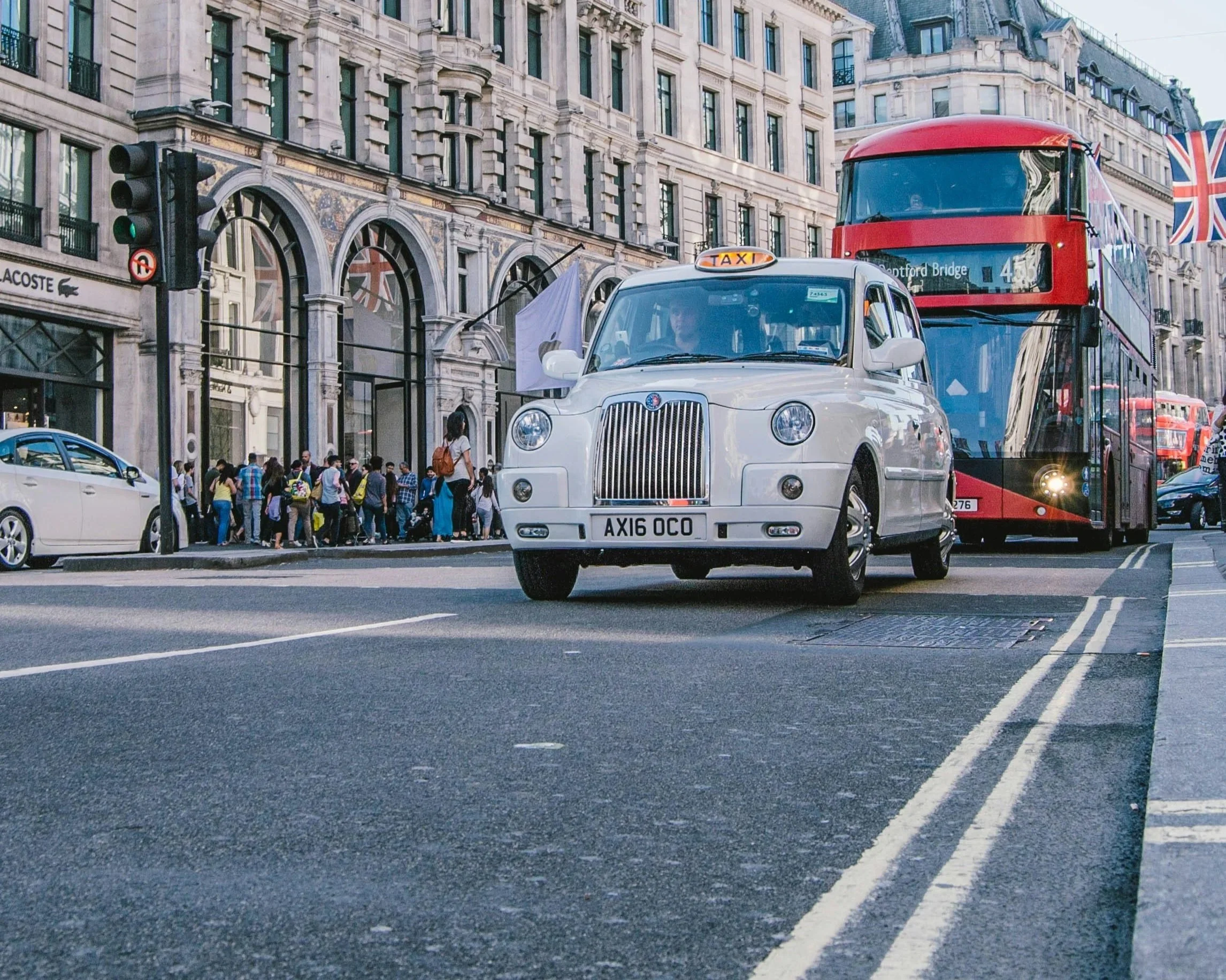 A white taxi cab and a red double-decker bus on a city street in London, with a crowd of pedestrians waiting to cross and buildings with Union Jack flags in the background.