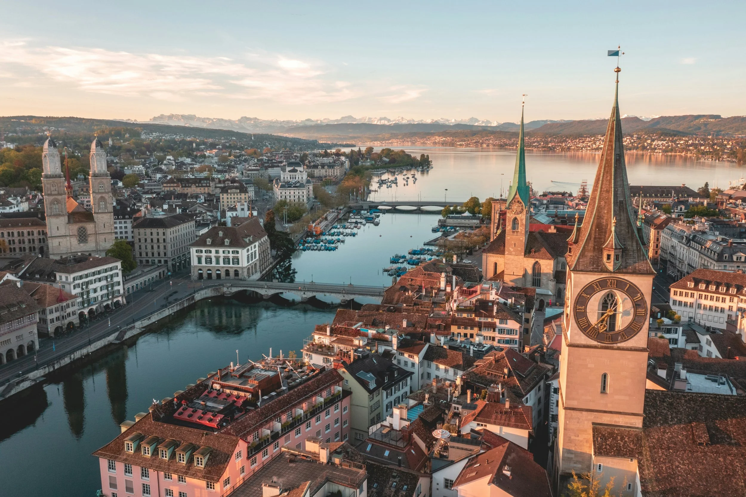 An aerial view of Zurich, Switzerland, featuring a large church with a tall clock tower, a river with bridges and boats, and a cityscape with historic buildings, mountains in the background, and a lake reflecting the sky at sunset.