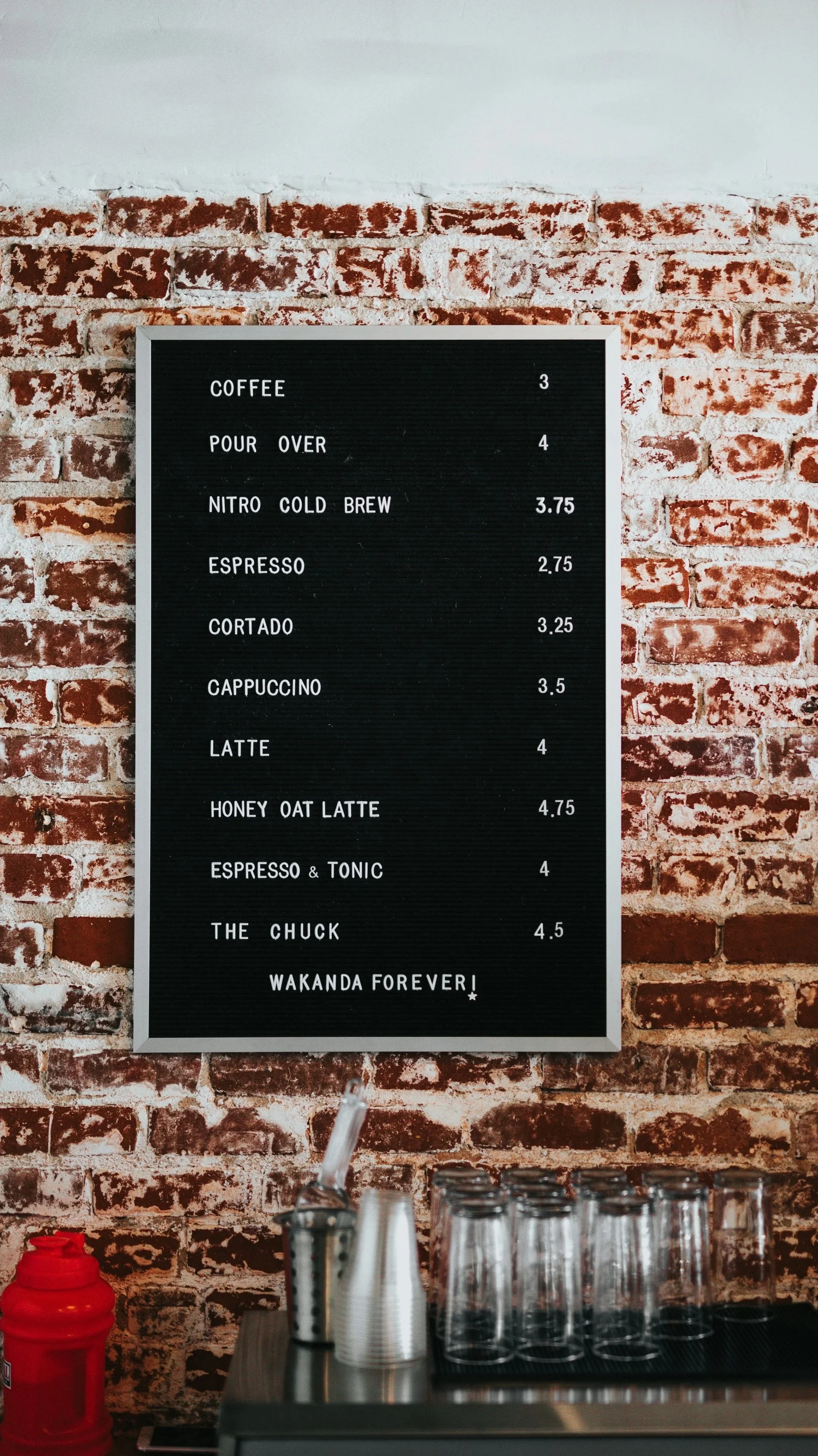 Coffee menu board with various coffee options and prices, positioned on a brick wall background, with cups and utensils below.