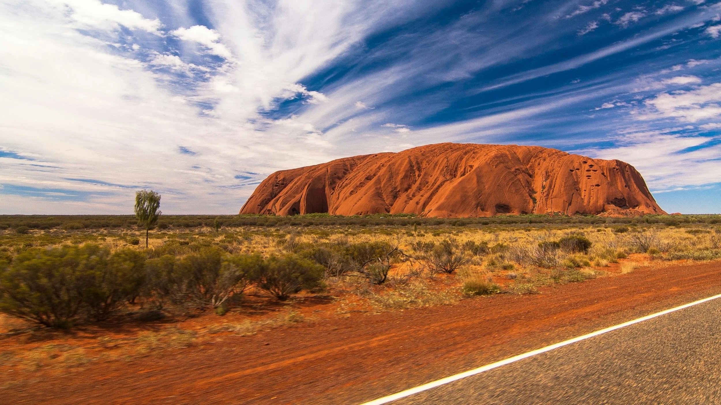 A large red sandstone formation called Uluru or Ayers Rock in the Australian desert, with a mostly clear sky and few clouds, and dry desert vegetation in the foreground along a paved road.
