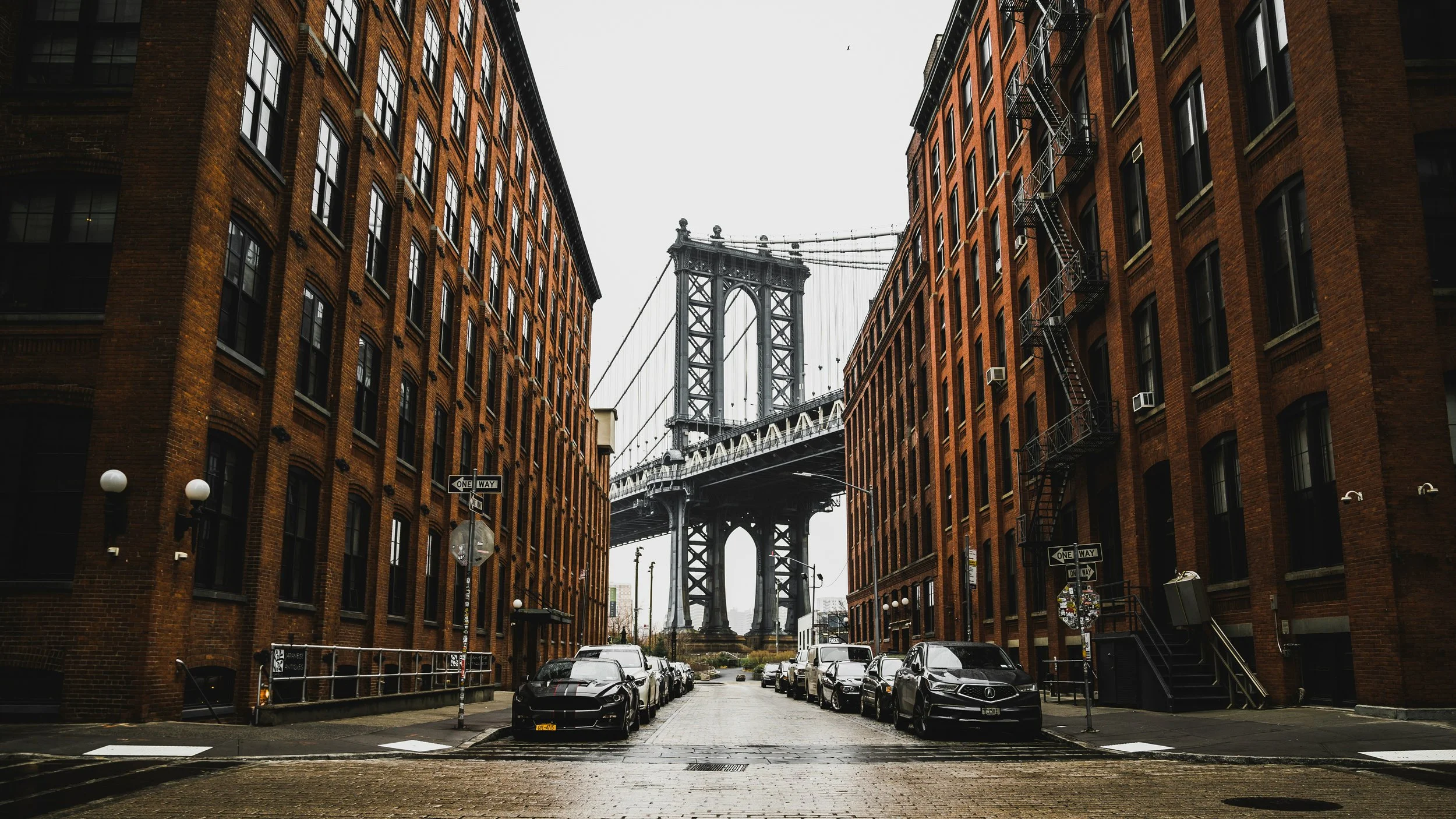 View of Manhattan Bridge framed by two red brick buildings on a cobblestone street with parked cars and street signs.