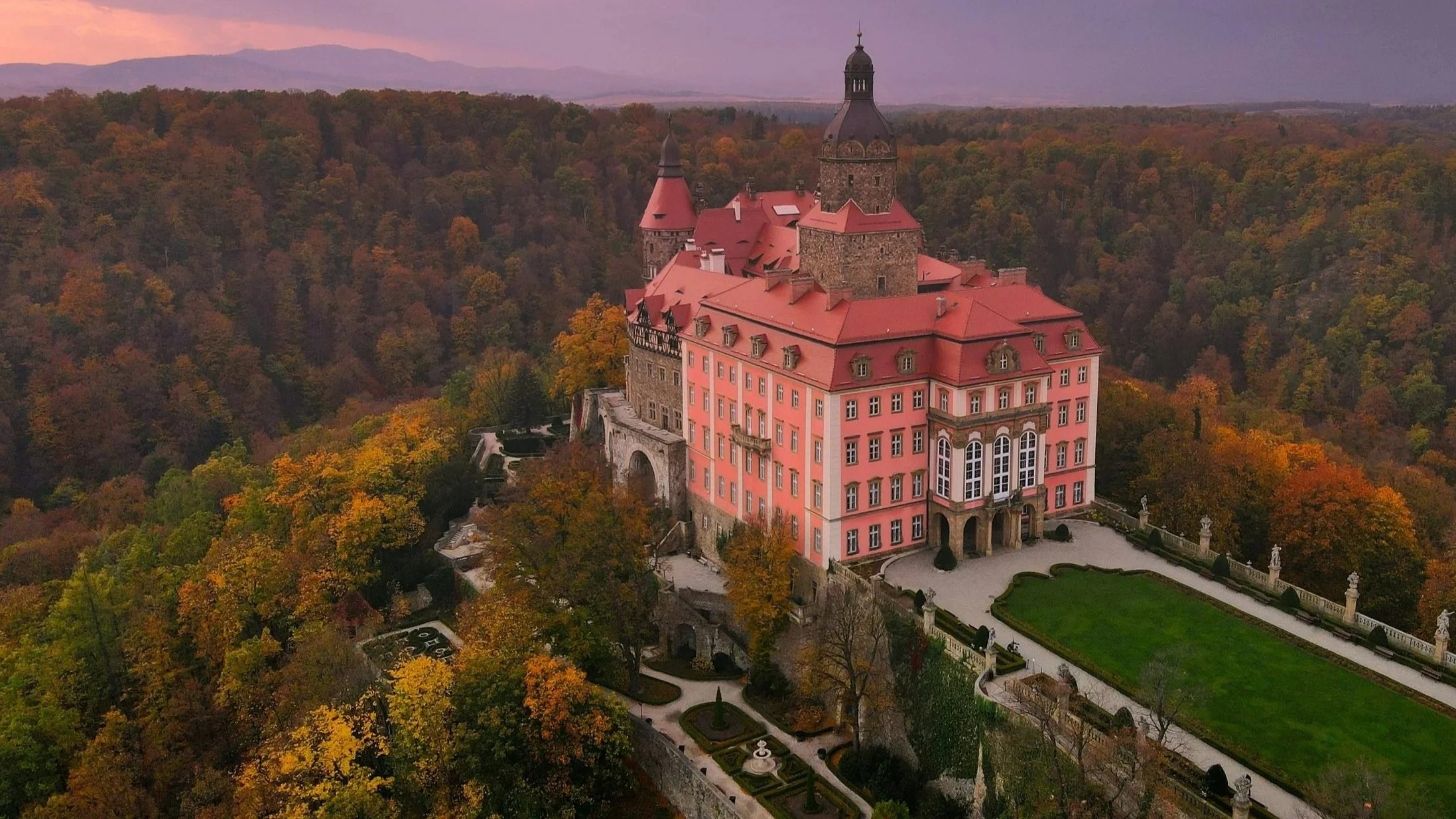 A medieval castle with pink walls and red roofs, surrounded by fall foliage, situated on a hill with landscaped gardens and pathways.