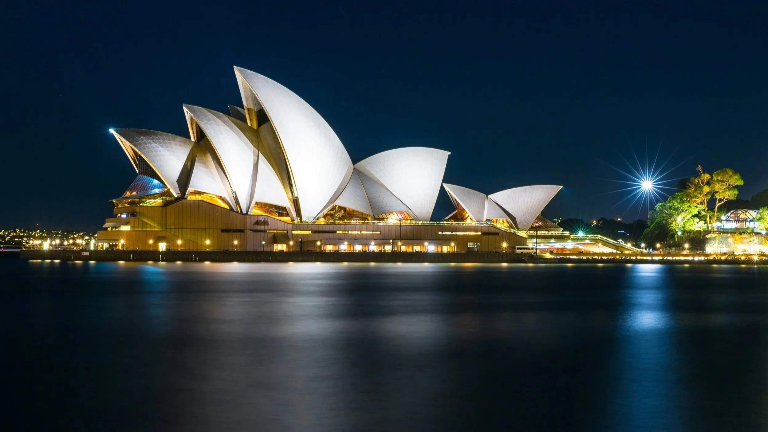 Night view of the Sydney Opera House illuminated along the waterfront with reflections in the water.