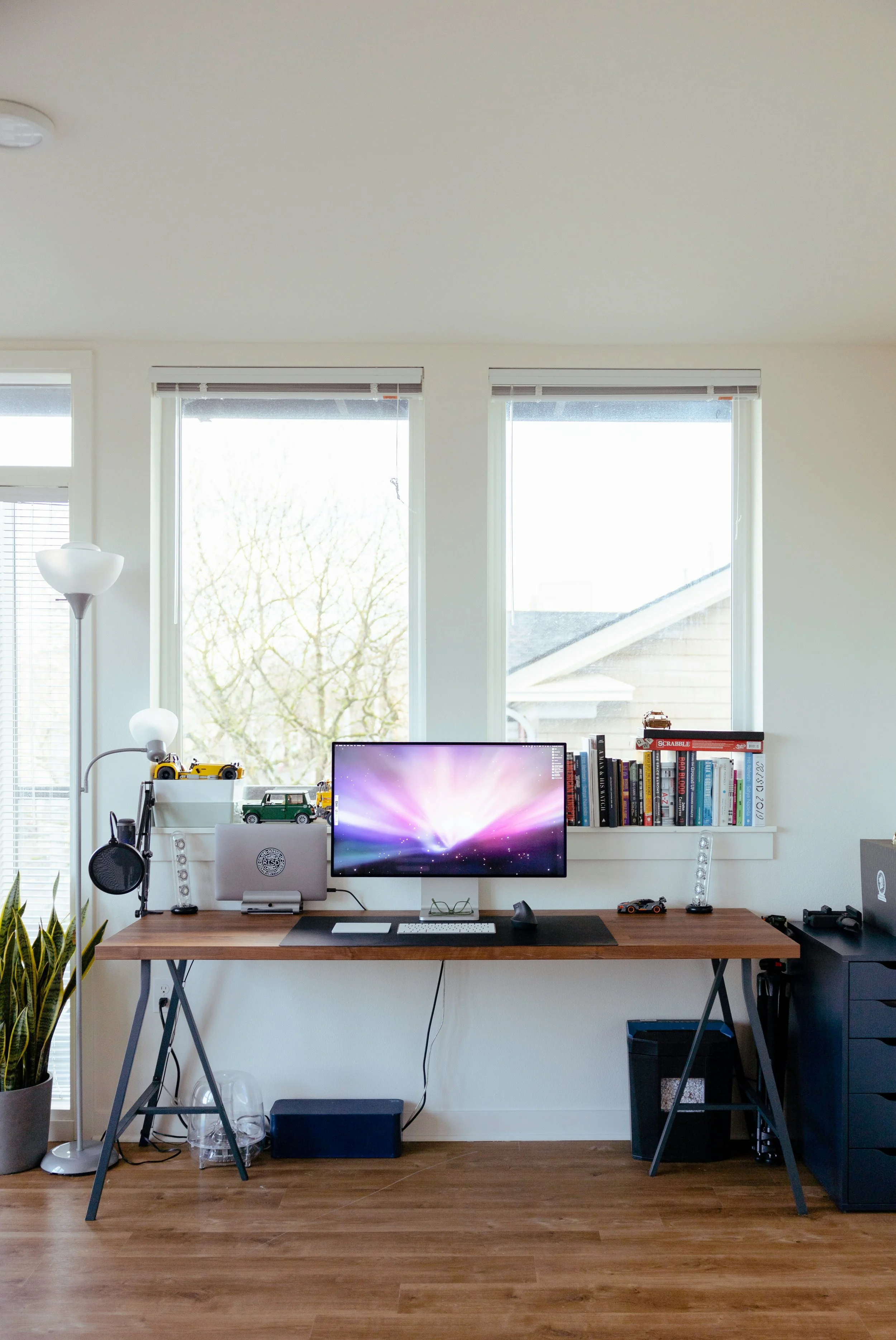 A home office setup with a wooden desk, a computer monitor, bookshelves, a laptop, and decorative items in front of three windows letting in natural light.