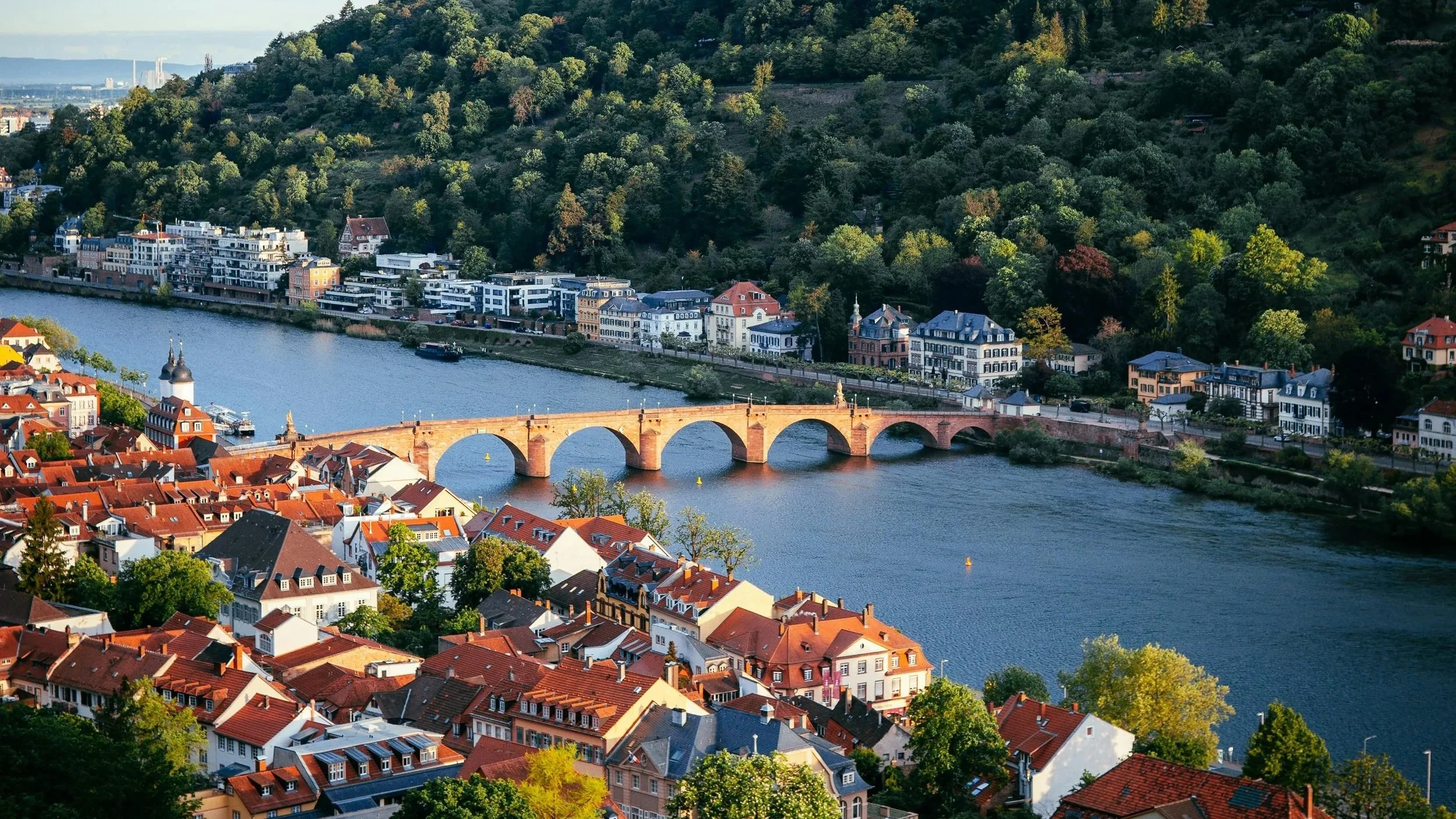 Aerial view of a city on a river with a historic brick bridge, colorful building rooftops, and green hills in the background.
