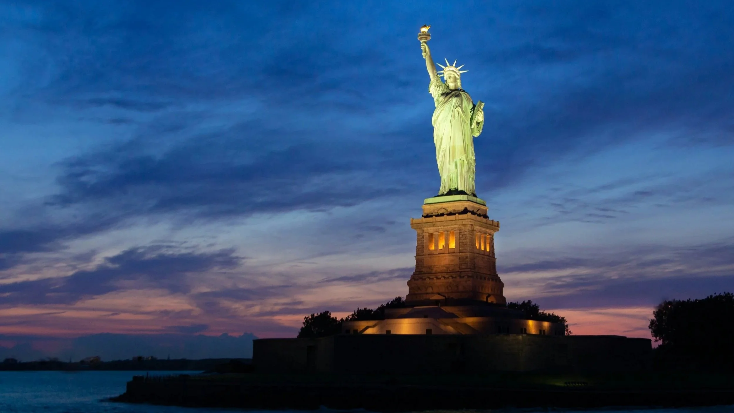 The Statue of Liberty illuminated at dusk with a colorful sky in background.