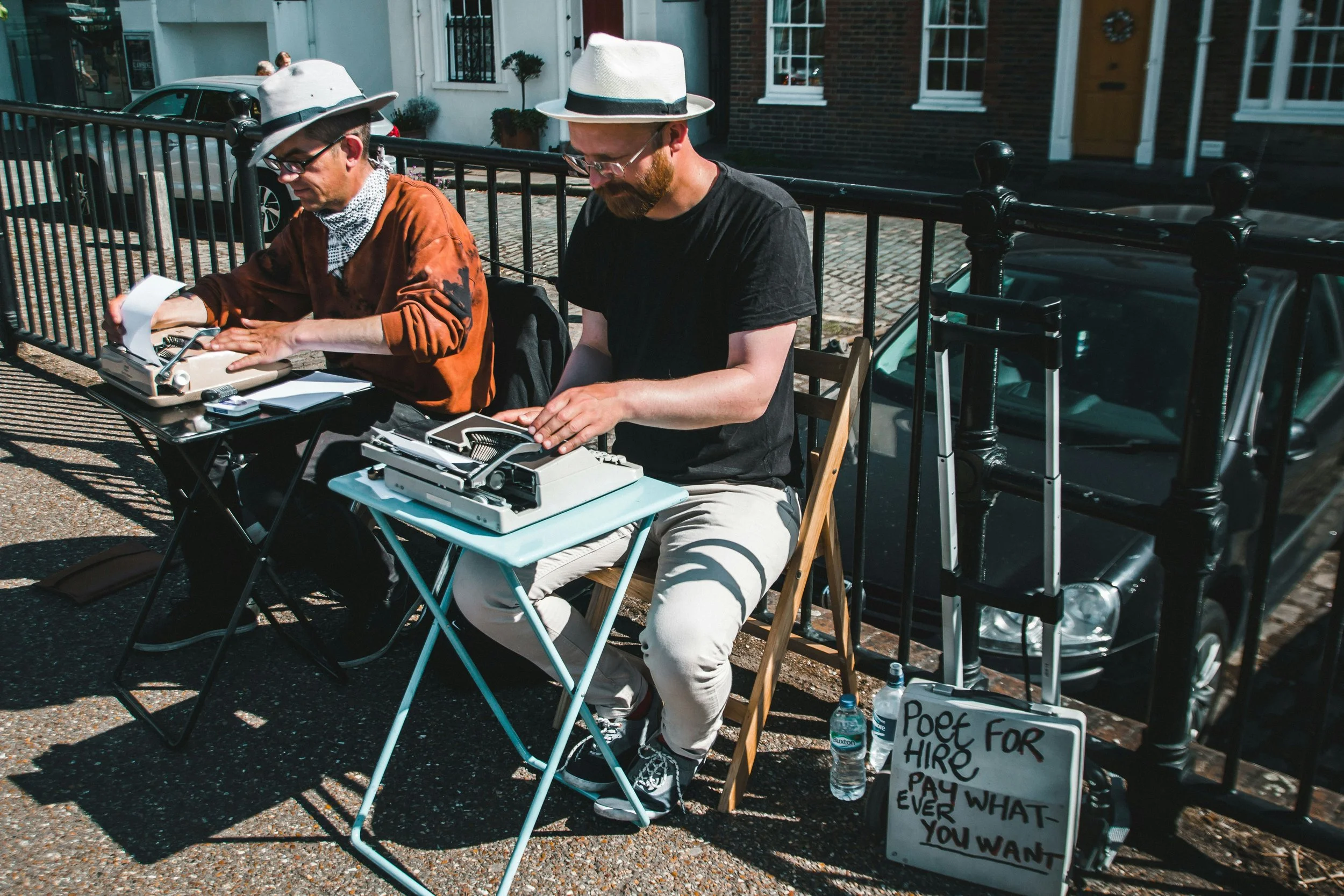 Two men sit at small tables on the sidewalk, typing on vintage typewriters. One wears a white hat, glasses, and an orange shirt; the other wears a white hat, glasses, and a black t-shirt. There is a whiteboard sign that says, 'Poet for hire pay what ever you want,' a water bottle, and a small ladder next to them.
