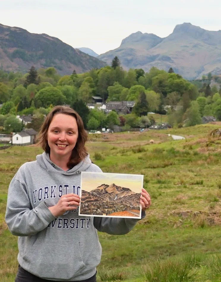 A woman in a light gray hoodie smiling outdoors in a green landscape, holding a painting of a mountain range. Behind her, there are trees, houses, and mountains in the distance.
