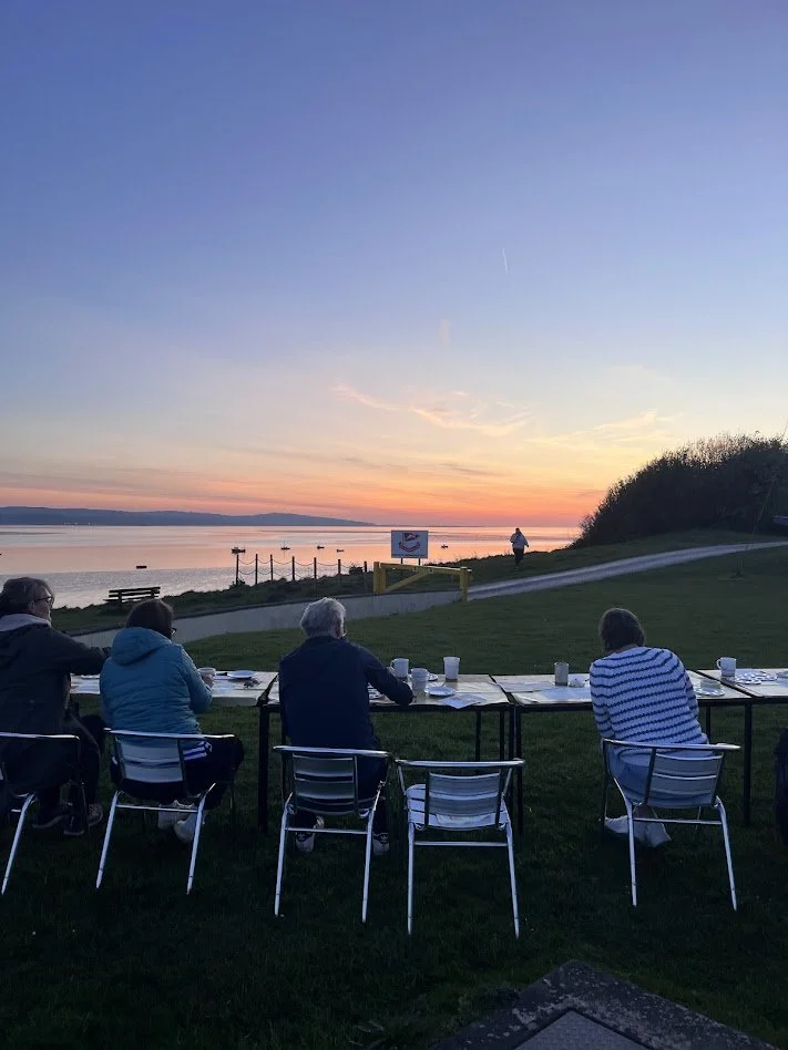 People dining outdoors by a lake during sunset, with a person walking on a path and boats on the water in the background.