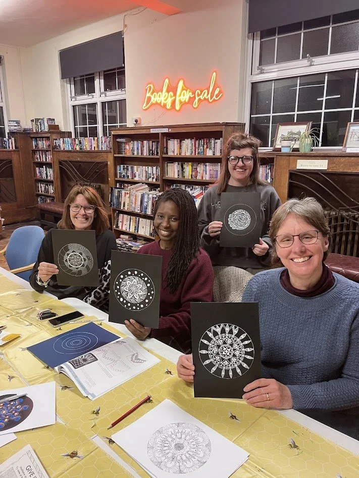 Four women sitting at a table in a library, holding up their paper designs with geometric patterns, with bookshelves and a 'Books for Sale' sign in the background.