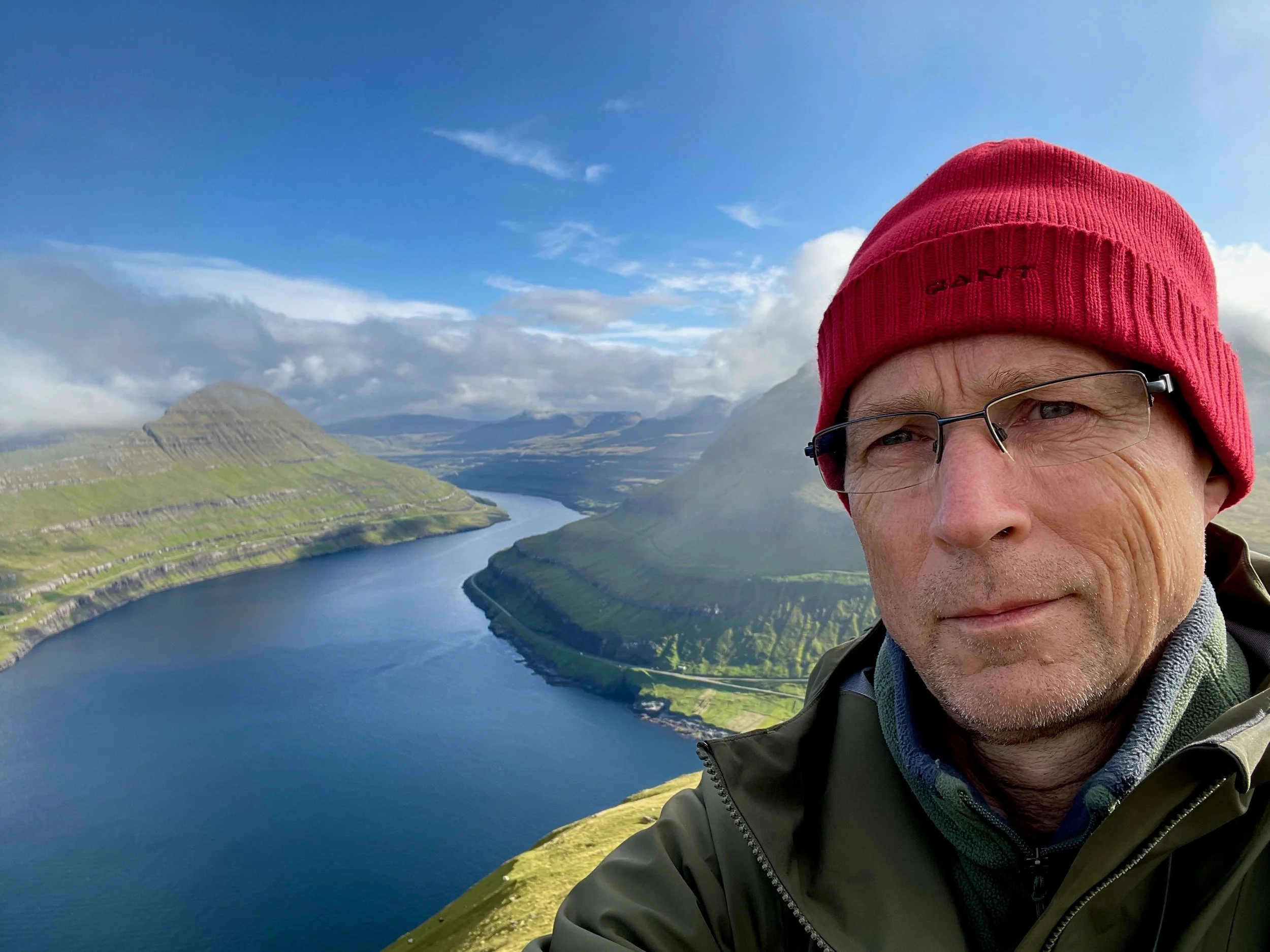 A man wearing a red beanie and glasses taking a selfie on a hill with a river and green mountains in the background.