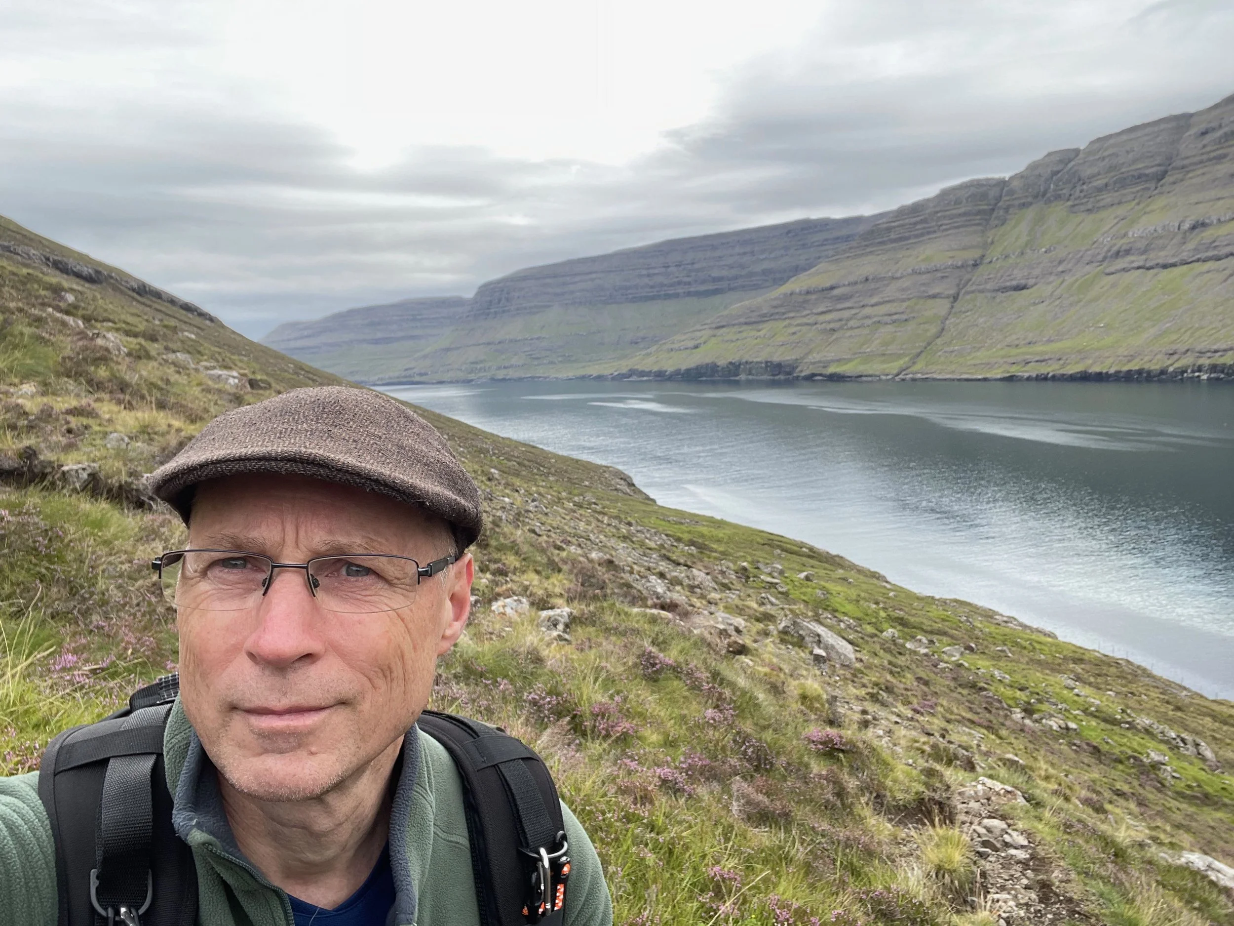 A man wearing glasses, a brown cap, and a green jacket takes a selfie outdoors with a river and grassy hills in the background.