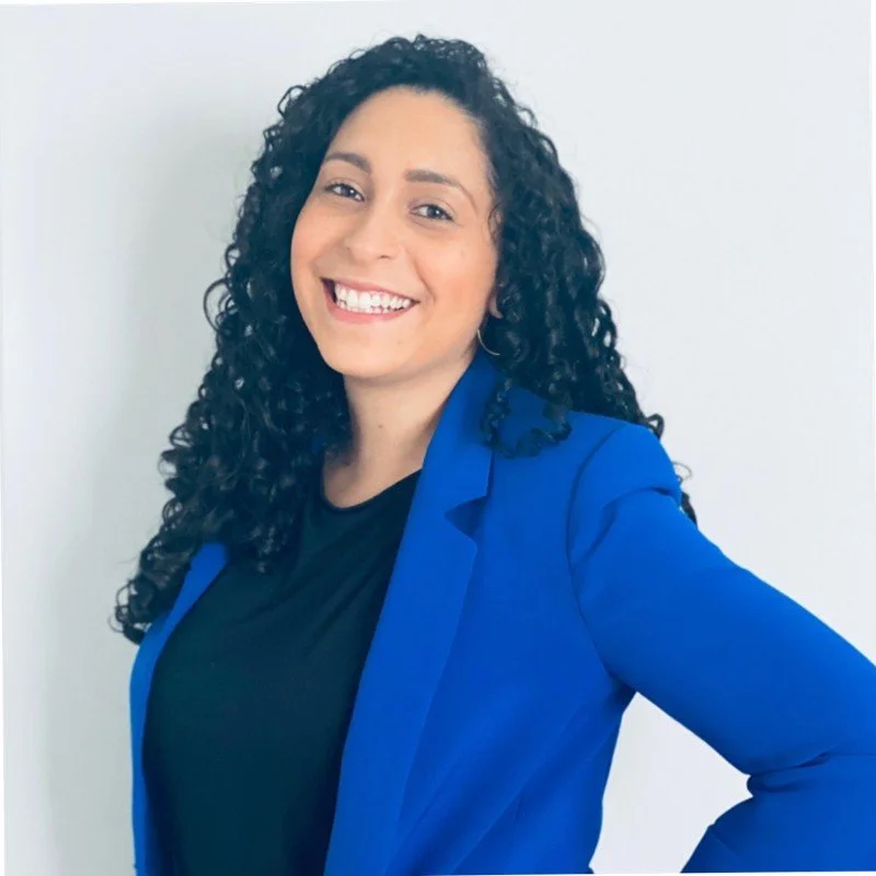 A woman with curly black hair smiling, wearing a blue blazer and black top, standing against a white background.