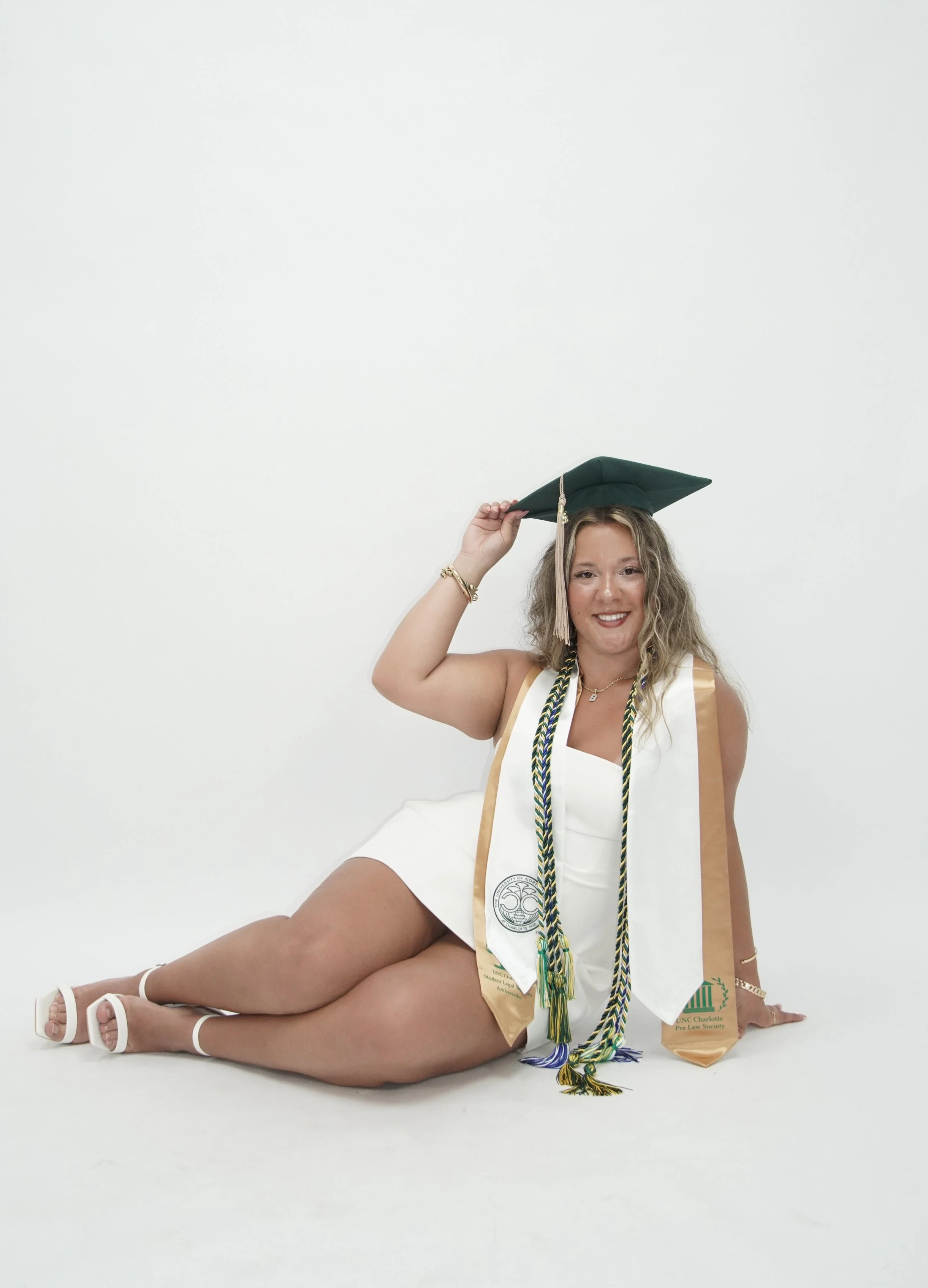 Woman in a white dress, wearing a graduation cap and cords, sitting with one leg bent, smiling against a white background.