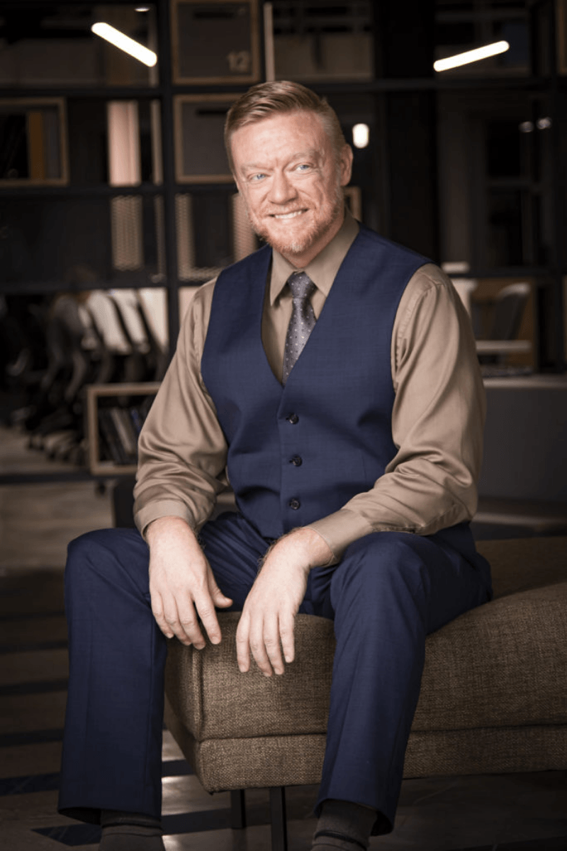 A man with red hair and a beard wearing a beige shirt, blue vest, and tie, sitting on a brown ottoman in a modern office space with dark walls and shelves.