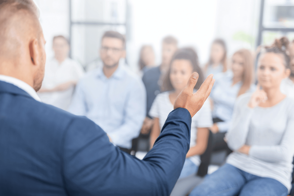 Person giving a presentation or speech to a group of audience members sitting in a room with natural lighting.