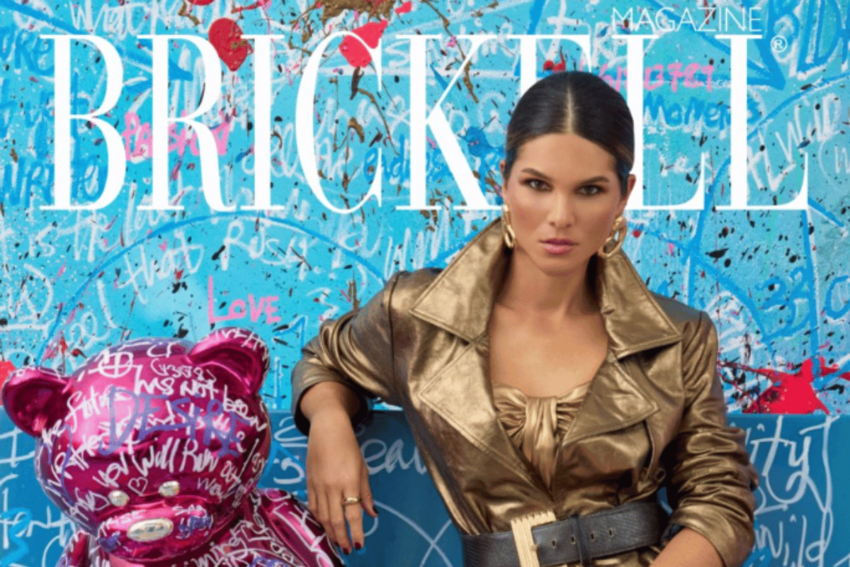 Cover of BRICKELL Magazine featuring a woman with dark hair in a gold jacket and large hoop earrings, sitting next to a shiny pink teddy bear balloon with white scribbled words, set against a colorful graffiti background.