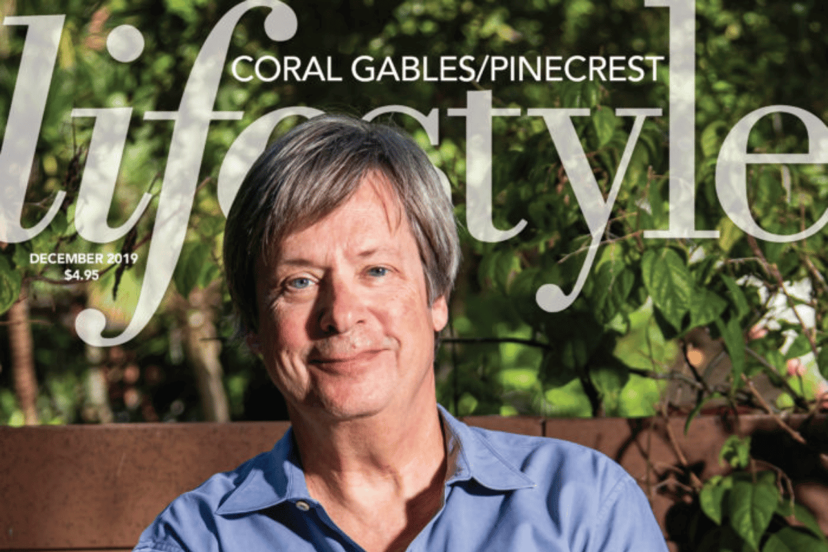 A man with gray hair and blue eyes sitting outside in front of green foliage, on the cover of Joyce magazine, December 2019, with a headline about coral gables/pinecrest lifestyle.