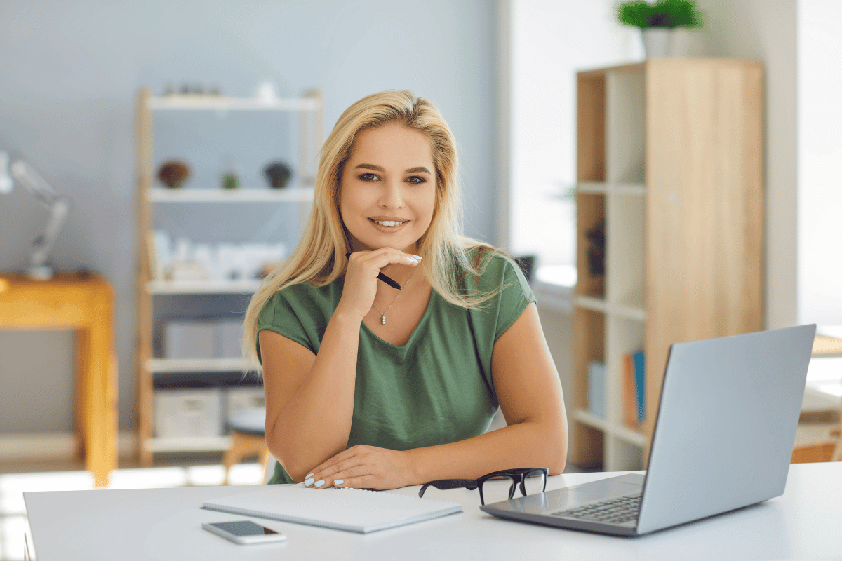 Young woman sitting at a desk with a laptop, notebook, glasses, and smartphone in a well-lit, modern office.
